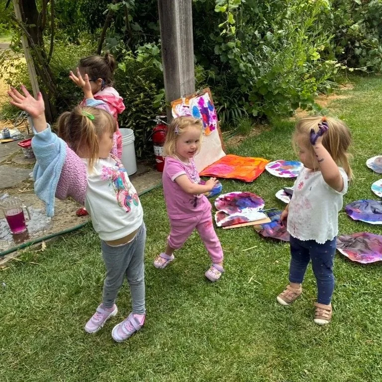 Four young girls are outdoors, engaging in a creative painting activity with colorful abstract paintings on in the grass area.