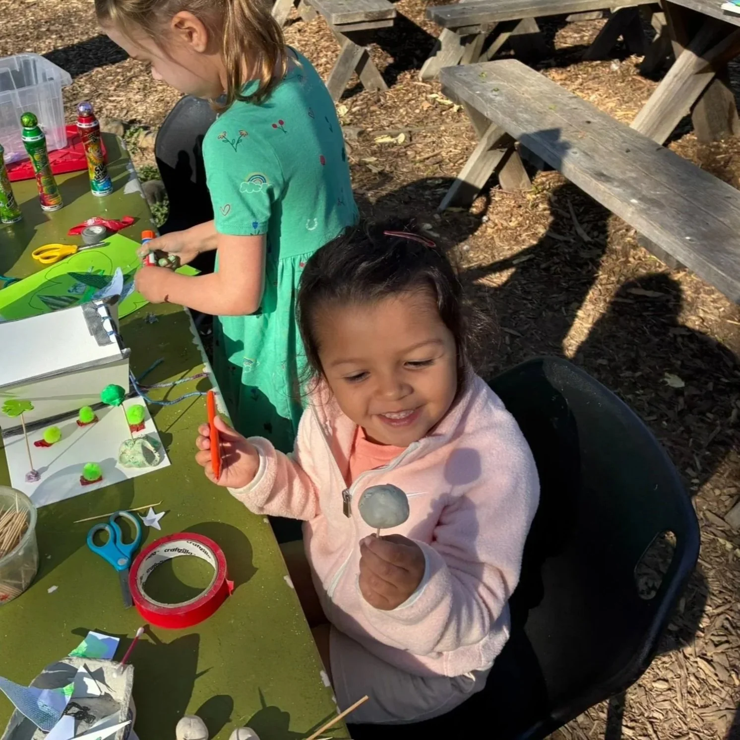 Two young girls sitting at an outdoor table making crafts with foam balls, sticks, and decorations on a green table, with tools such as scissors, tape, and glue visible. One girl is smiling and holding a craft item, while the other girl is focused on her crafts.