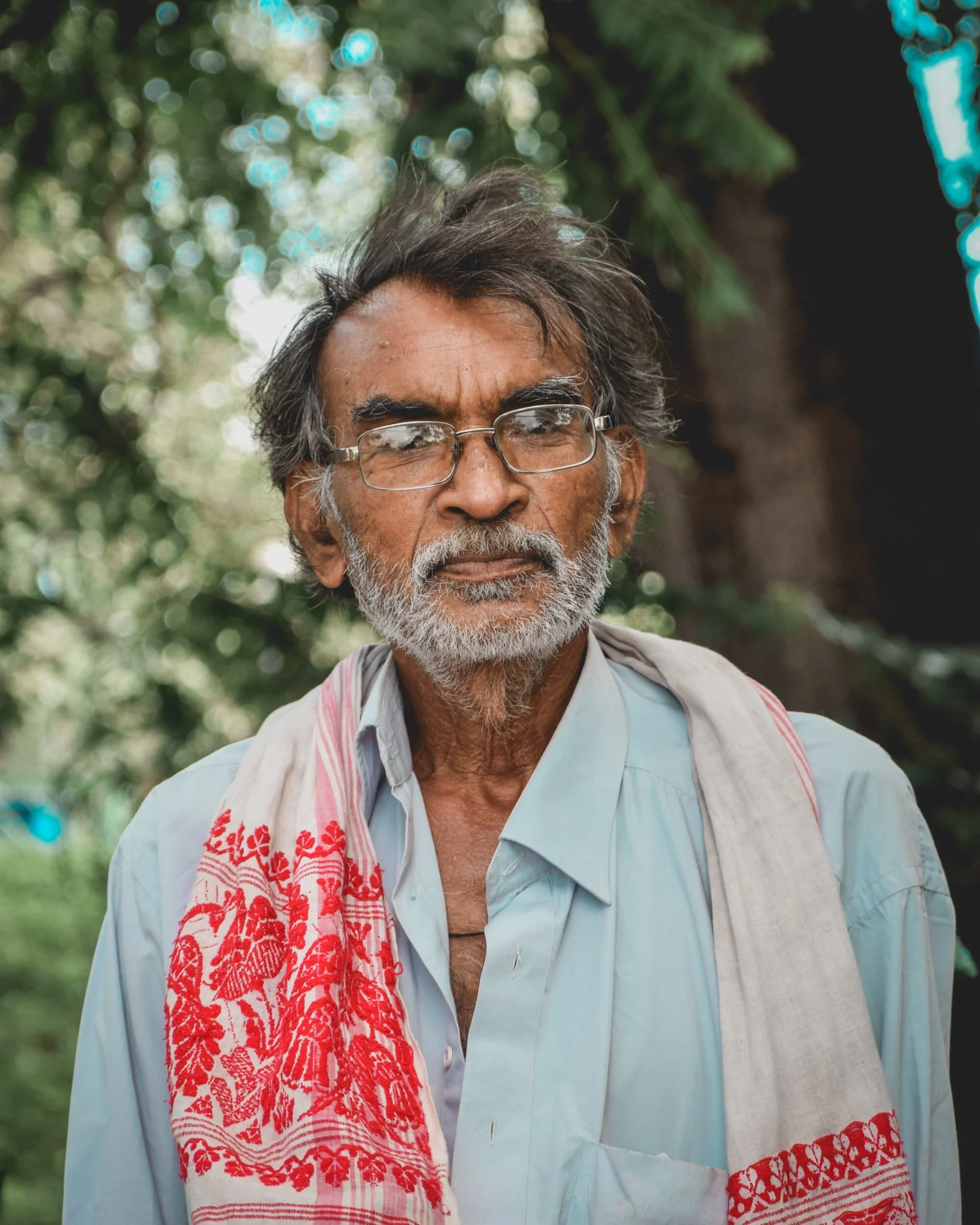An elderly man with glasses, gray hair, and a beard standing outdoors surrounded by greenery, wearing a light blue shirt and a beige cloth with red embroidery draped over his shoulders.