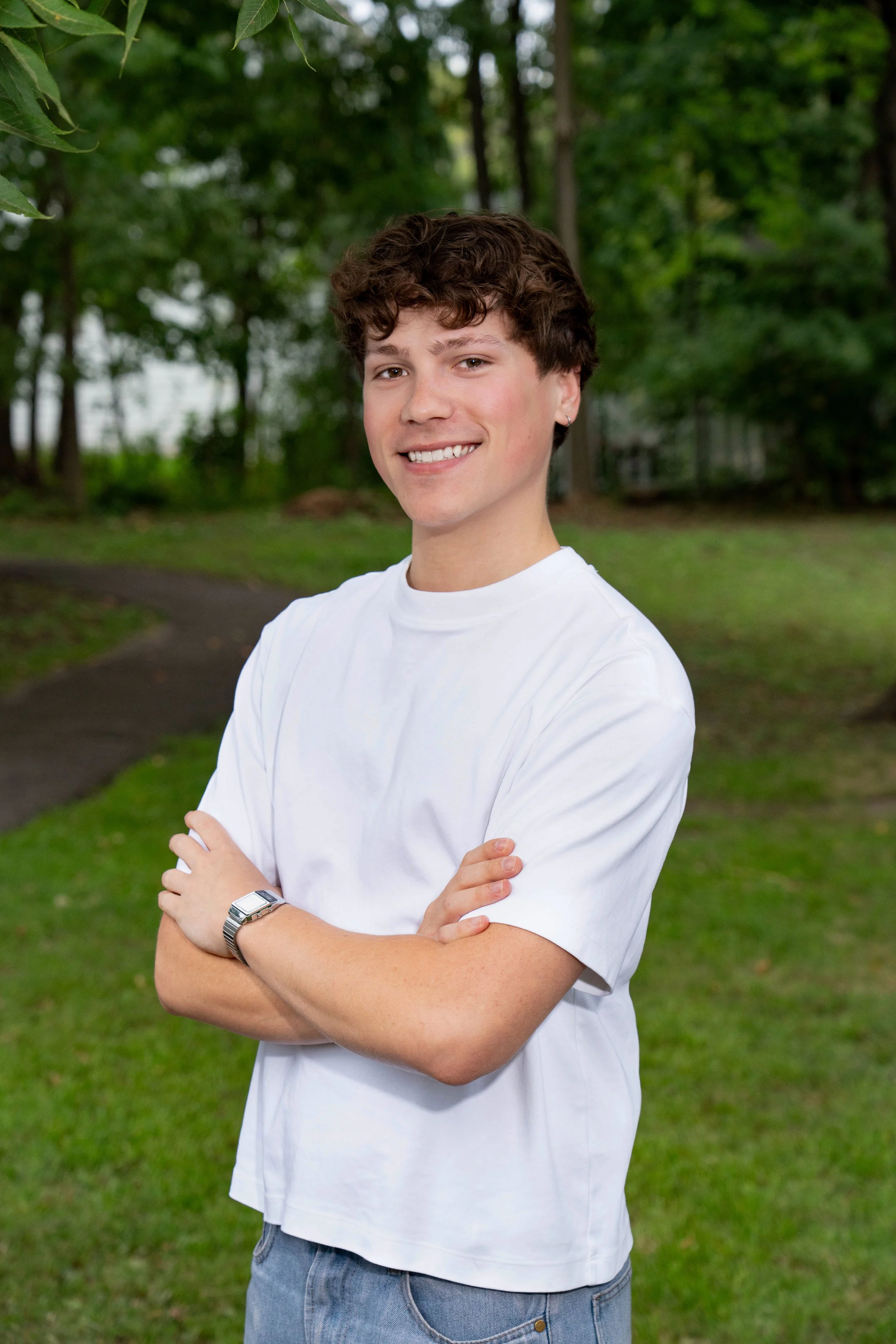 A young man with curly brown hair, wearing a white t-shirt and blue jeans, standing outdoors with his arms crossed, smiling, in front of a background of green trees and grass.