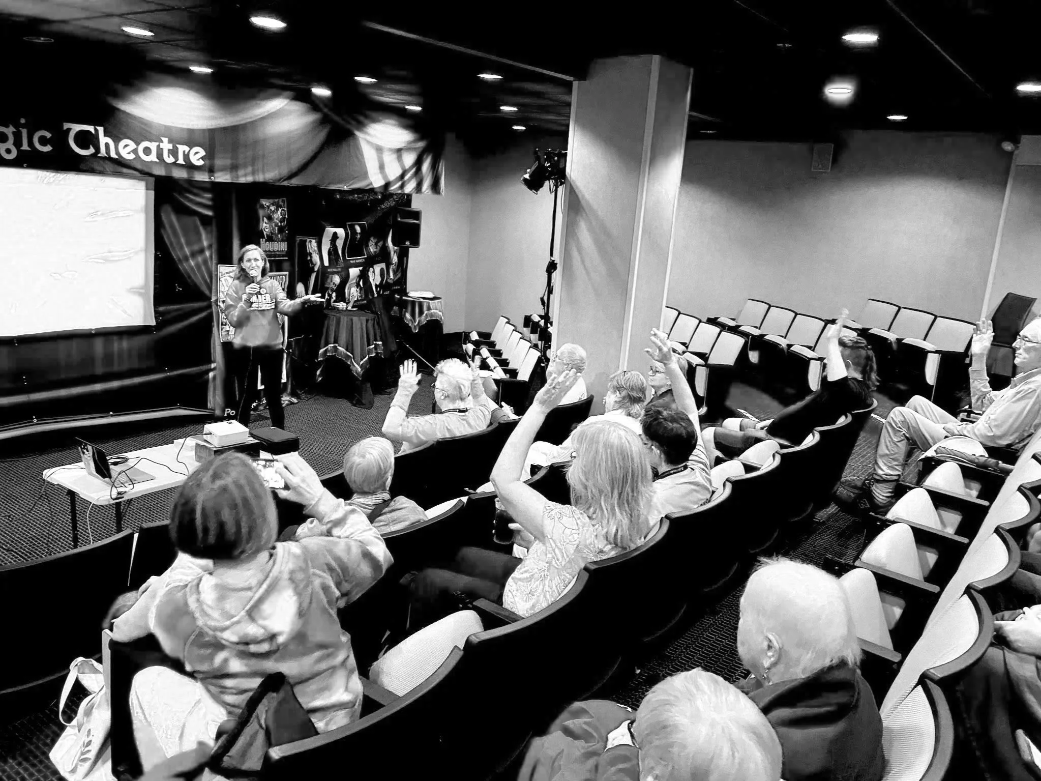 A presenter speaking in front of an audience in a small theater or conference room, with several people raising their hands to ask questions or participate.
