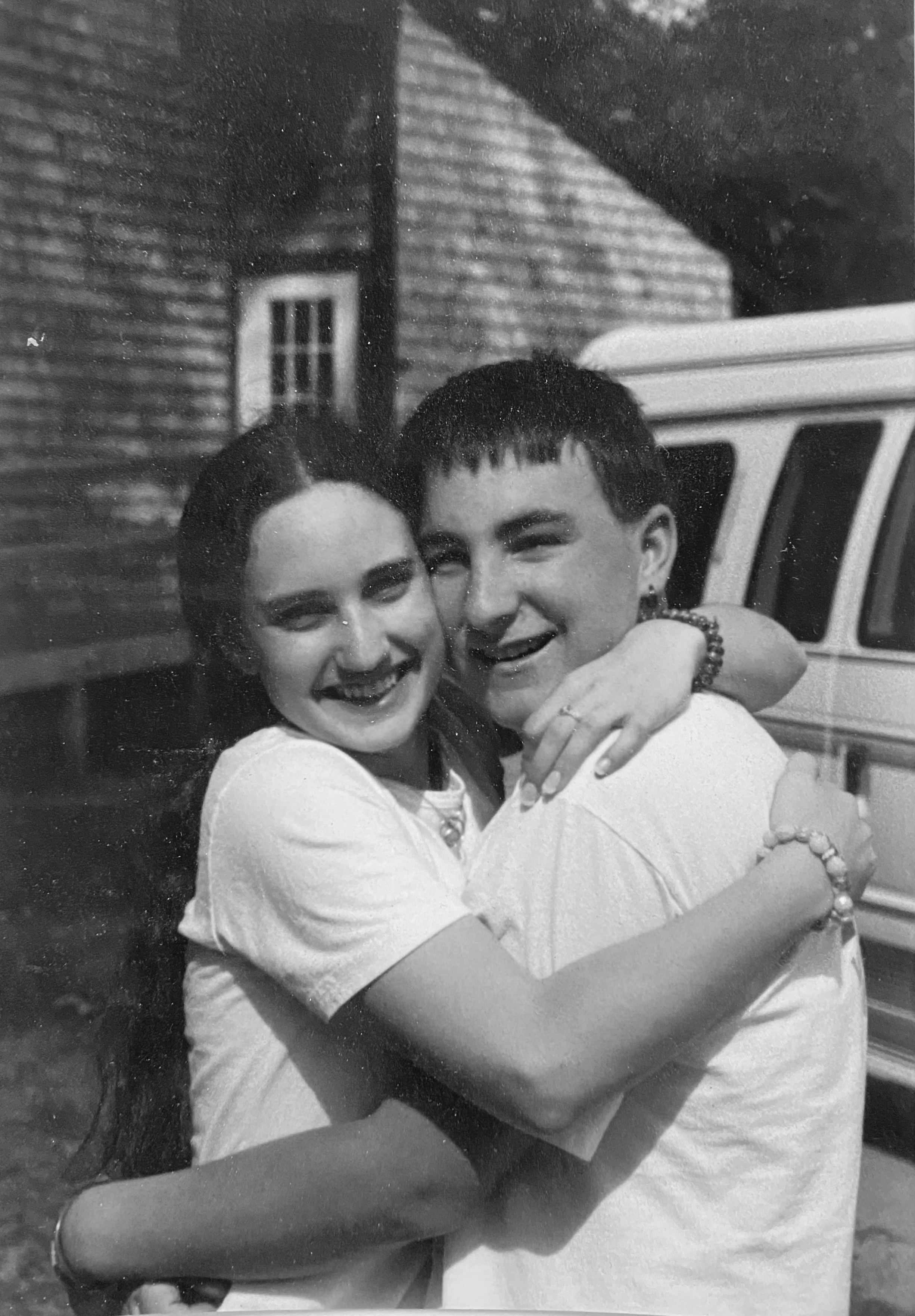 A young couple hugging, smiling, and looking happy in front of a house and a vehicle.