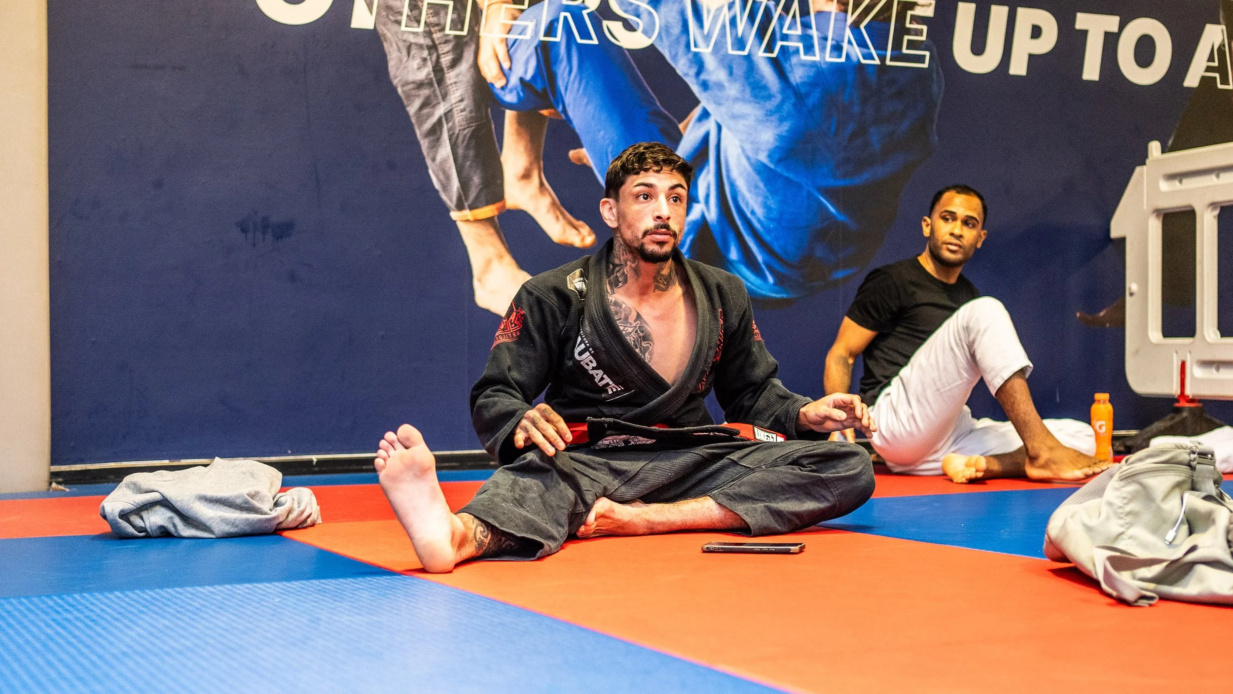 A male Brazilian Jiu-Jitsu practitioner sitting on a blue and red mat, wearing a black gi with a red belt, in a martial arts gym. Behind him, another person in a black t-shirt and white pants is seated on the mat. The background features a large post