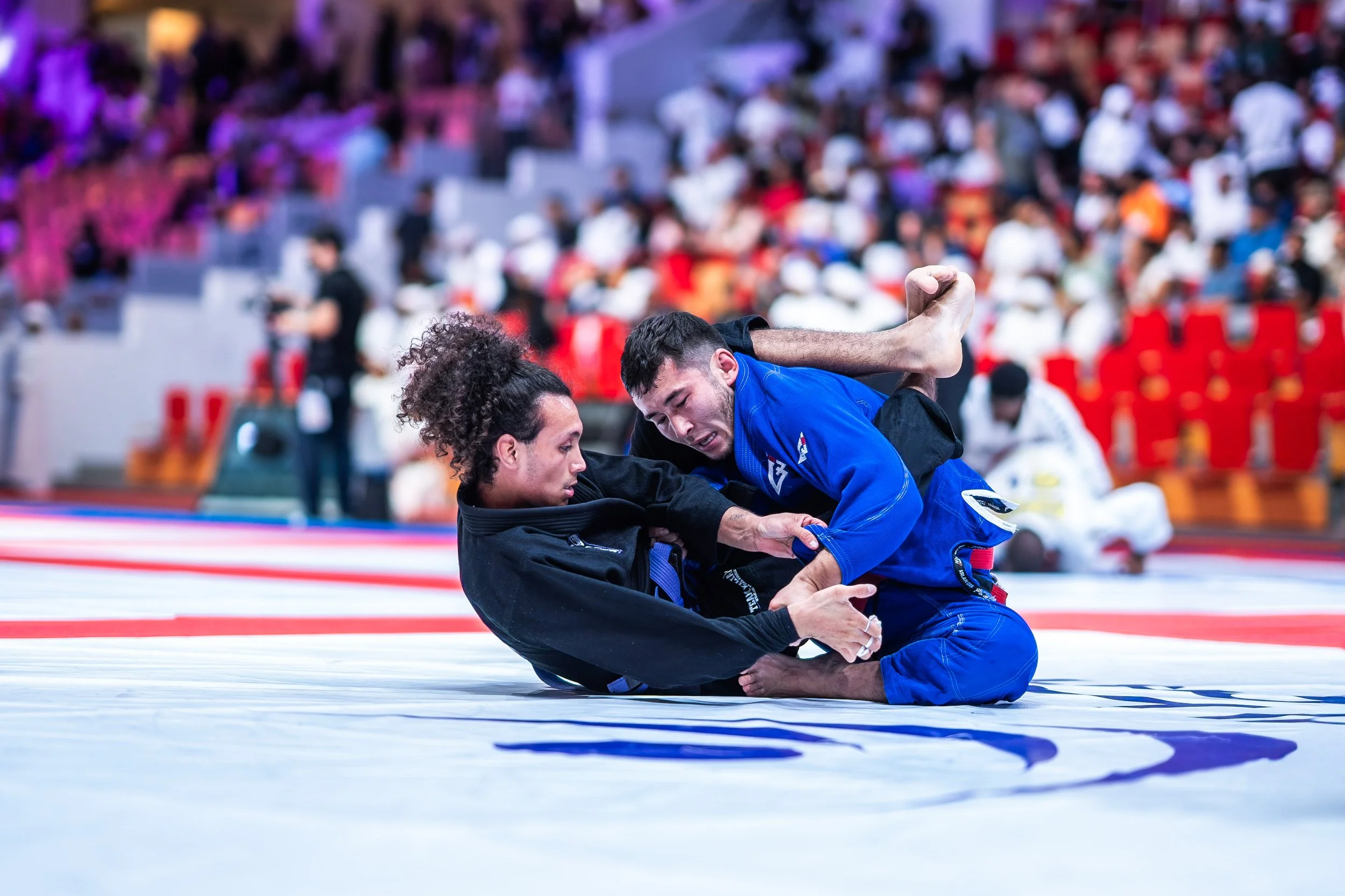 Two female Brazilian Jiu-Jitsu competitors grappling on a mat during a tournament, with one in a black gi and the other in a blue gi, surrounded by spectators.
