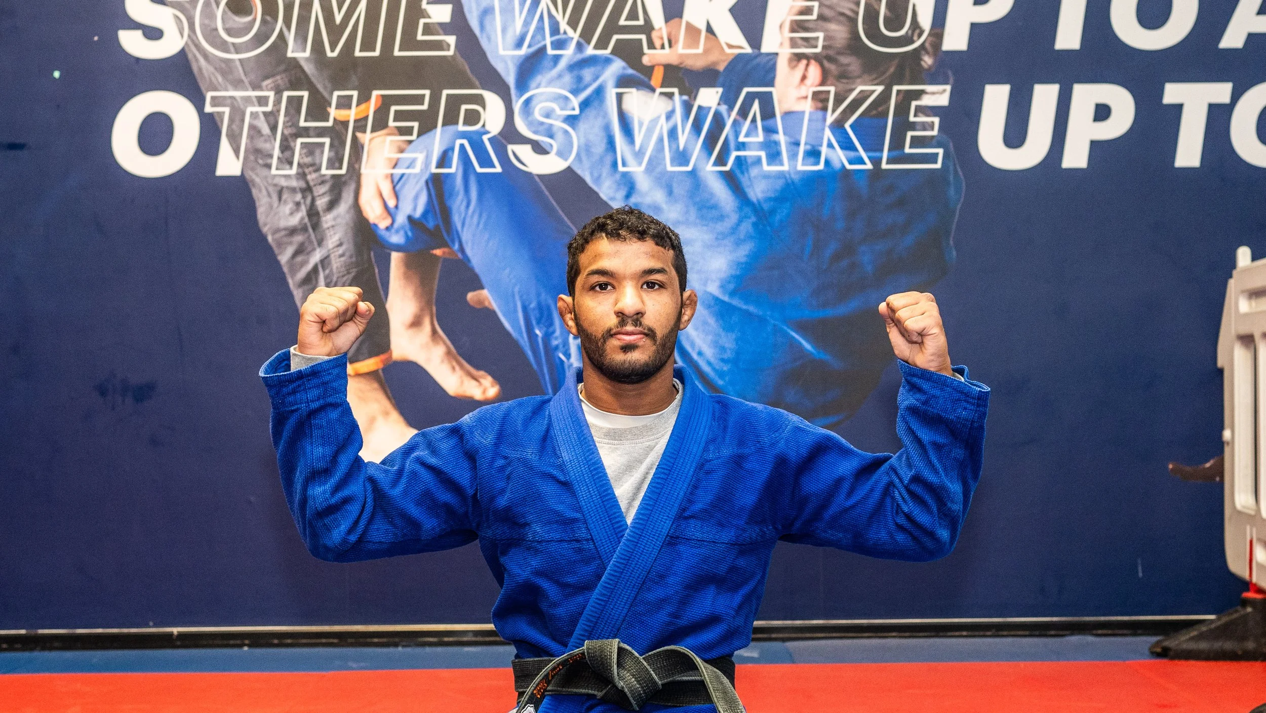 A man in a blue martial arts gi and black belt poses with flexed arms in front of a large banner with partially visible motivational text. The background includes images of people in martial arts uniforms.