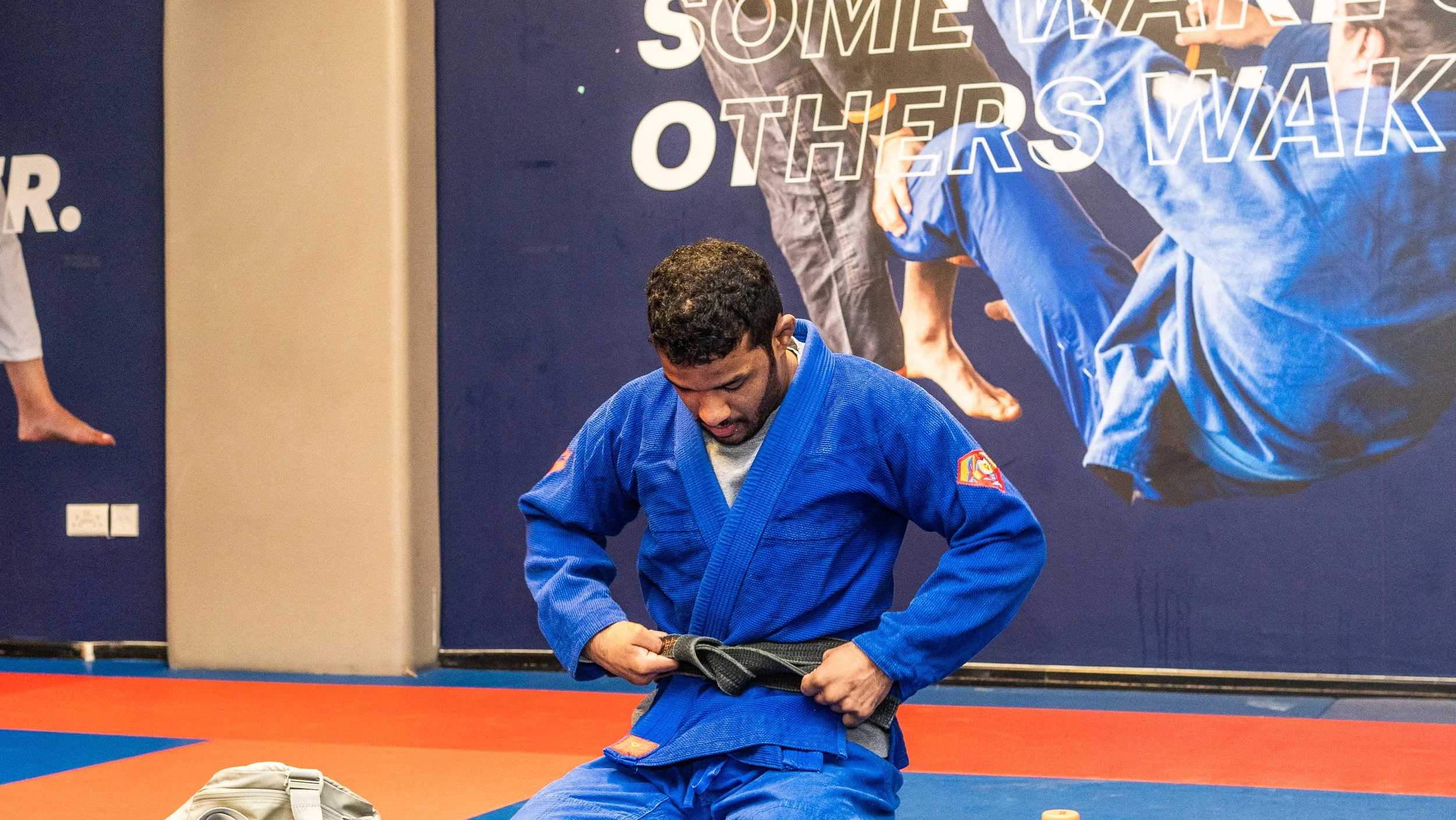 A man in a blue martial arts uniform, likely a Brazilian Jiu-Jitsu gi, is fastening his black belt in a martial arts training facility. A large poster with people in blue gi uniforms and the words 'SOME WARS OTHER WAK' is visible in the background, a