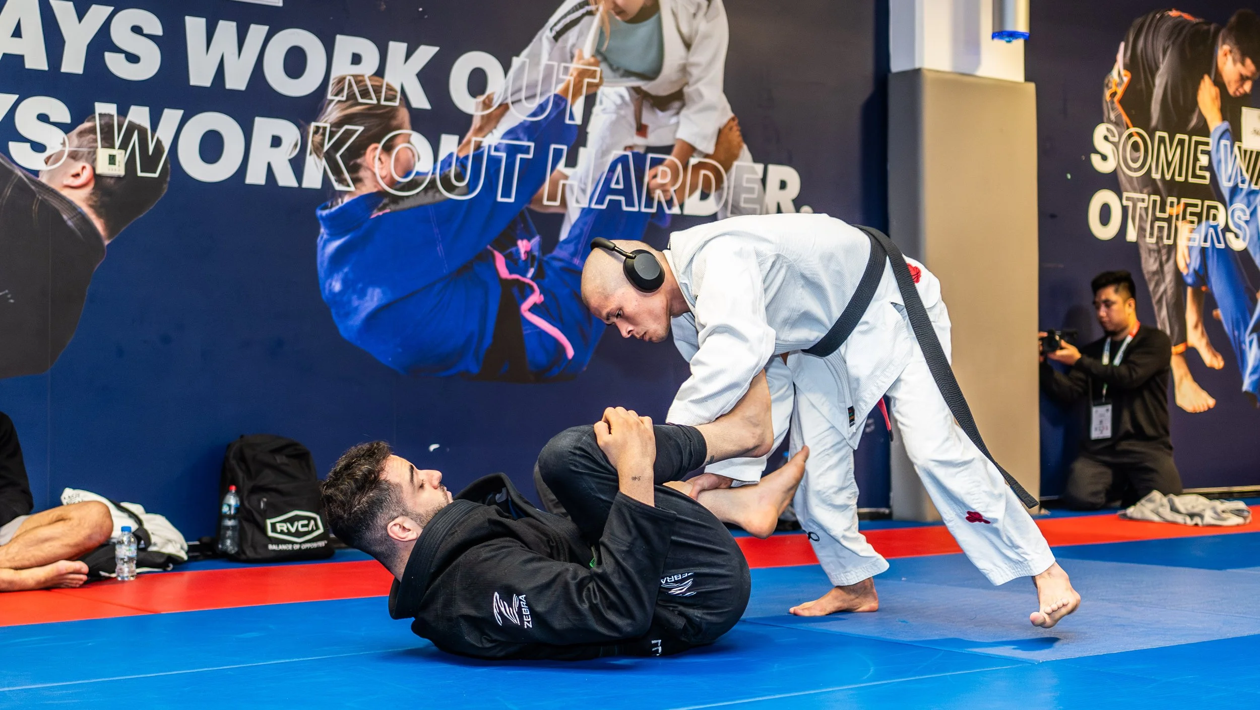 Two men practicing Brazilian Jiu-Jitsu on a blue mat. One man in a black gi is on his back, while the other man in a white gi is leaning over him, gripping his leg. A photographer is capturing the scene, and there are other individuals seated against