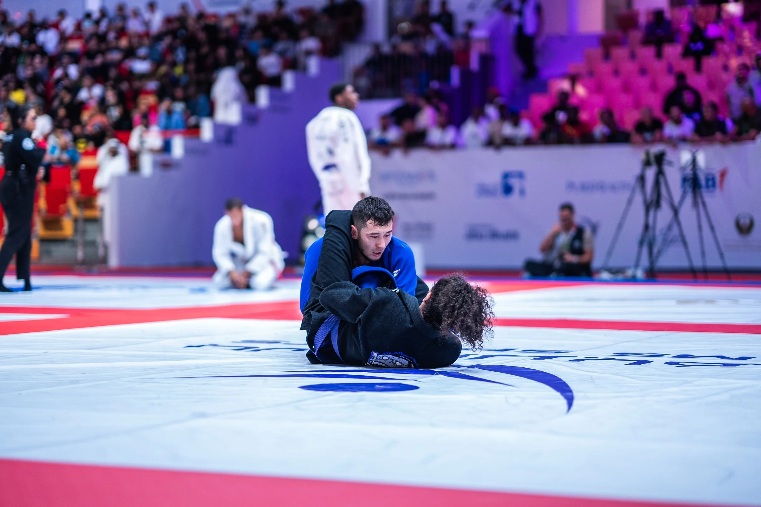 Two martial artists engaged in a Brazilian Jiu-Jitsu match on a mat, surrounded by spectators and officials, with one seated and the other attempting a submission move.
