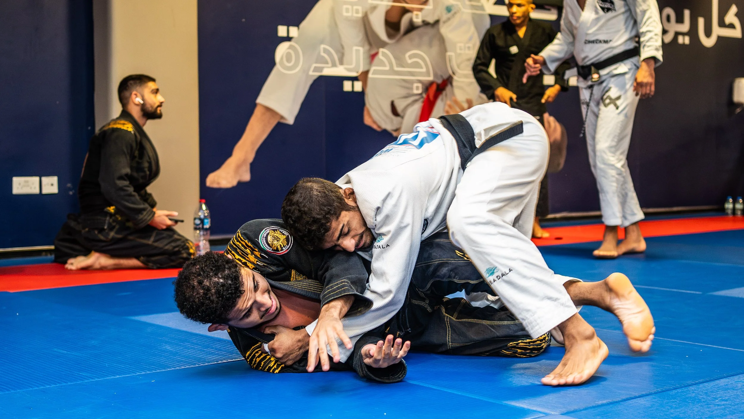 Two women practicing Brazilian Jiu-Jitsu on a blue mat, with one woman in a white gi on top, pinning the other woman in a black gi against the mat, while others watch in the background.