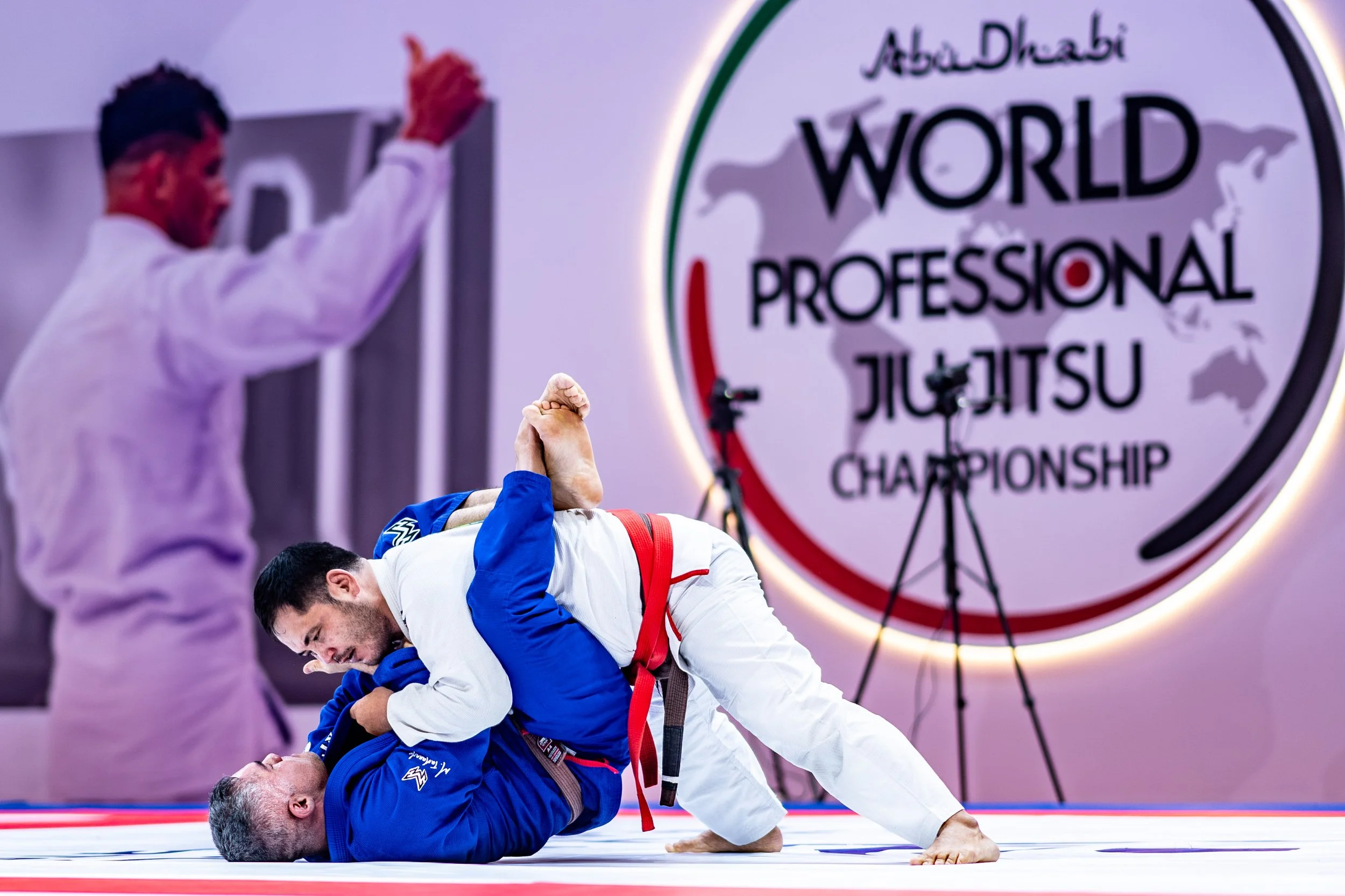 Two male jiu-jitsu competitors, one in blue and the other in white, grappling on the mat during an Abu Dhabi World Professional Jiu-Jitsu Championship. A large sign with the event's name is visible in the background.
