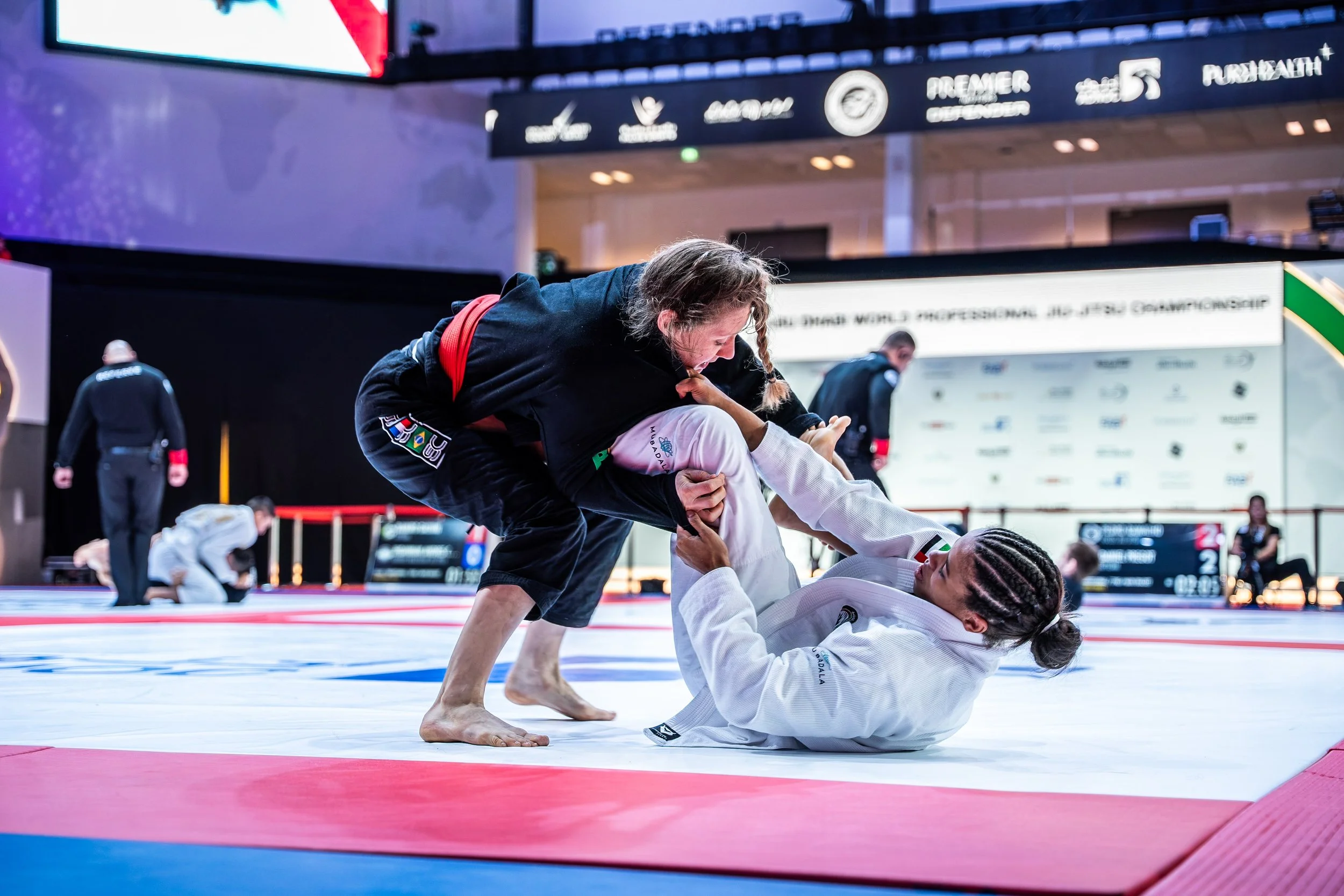 Two women competing in a Brazilian Jiu-Jitsu match on a mat, with one woman in a black gi and the other in a white gi. The woman in black is on top, gripping the collar of the woman in white, who is on her back with her legs wrapped around her oppone