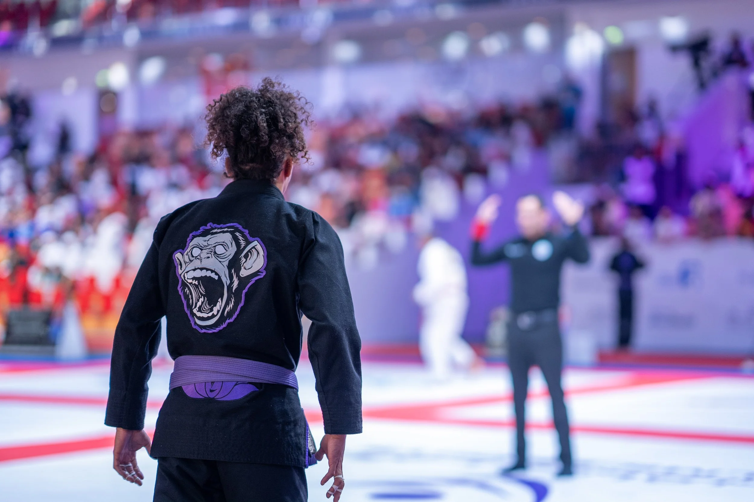 A person in a black Brazilian Jiu-Jitsu gi with a purple belt and a patch of a roaring ape face on the back, observing a match at a gym or tournament with blurred referees and spectators in the background.