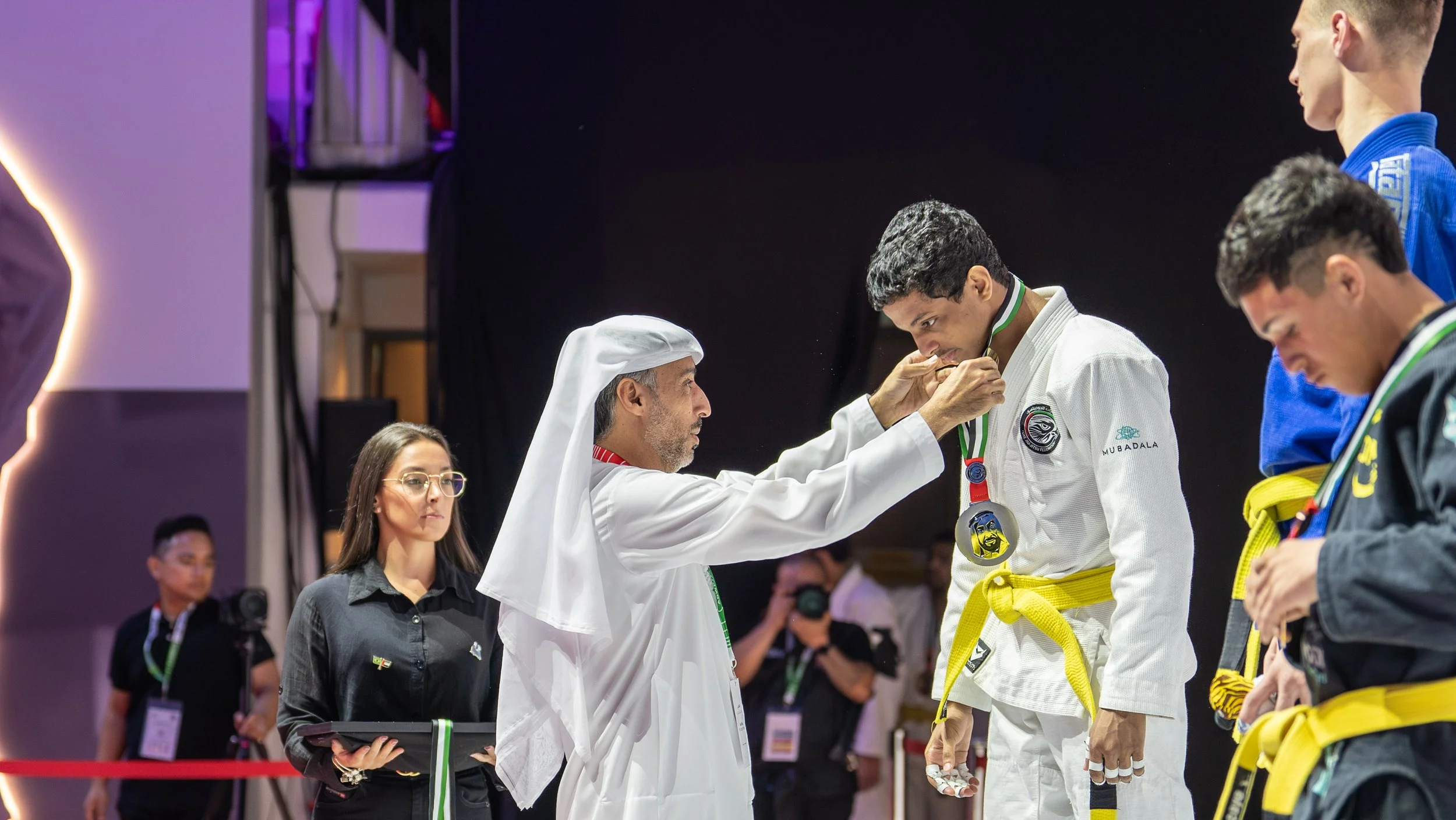 A man in traditional Middle Eastern attire awards a gold medal to a young male martial artist in a white judo gi, while other competitors and officials look on.