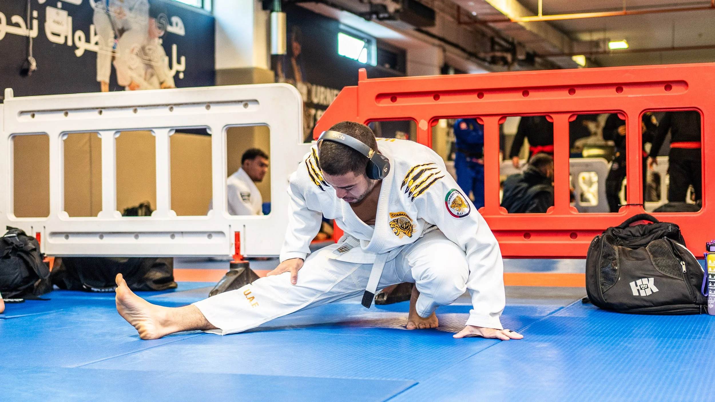 A man in a martial arts gi stretching on a blue mat at a martial arts training or competition venue, with other people and equipment in the background.