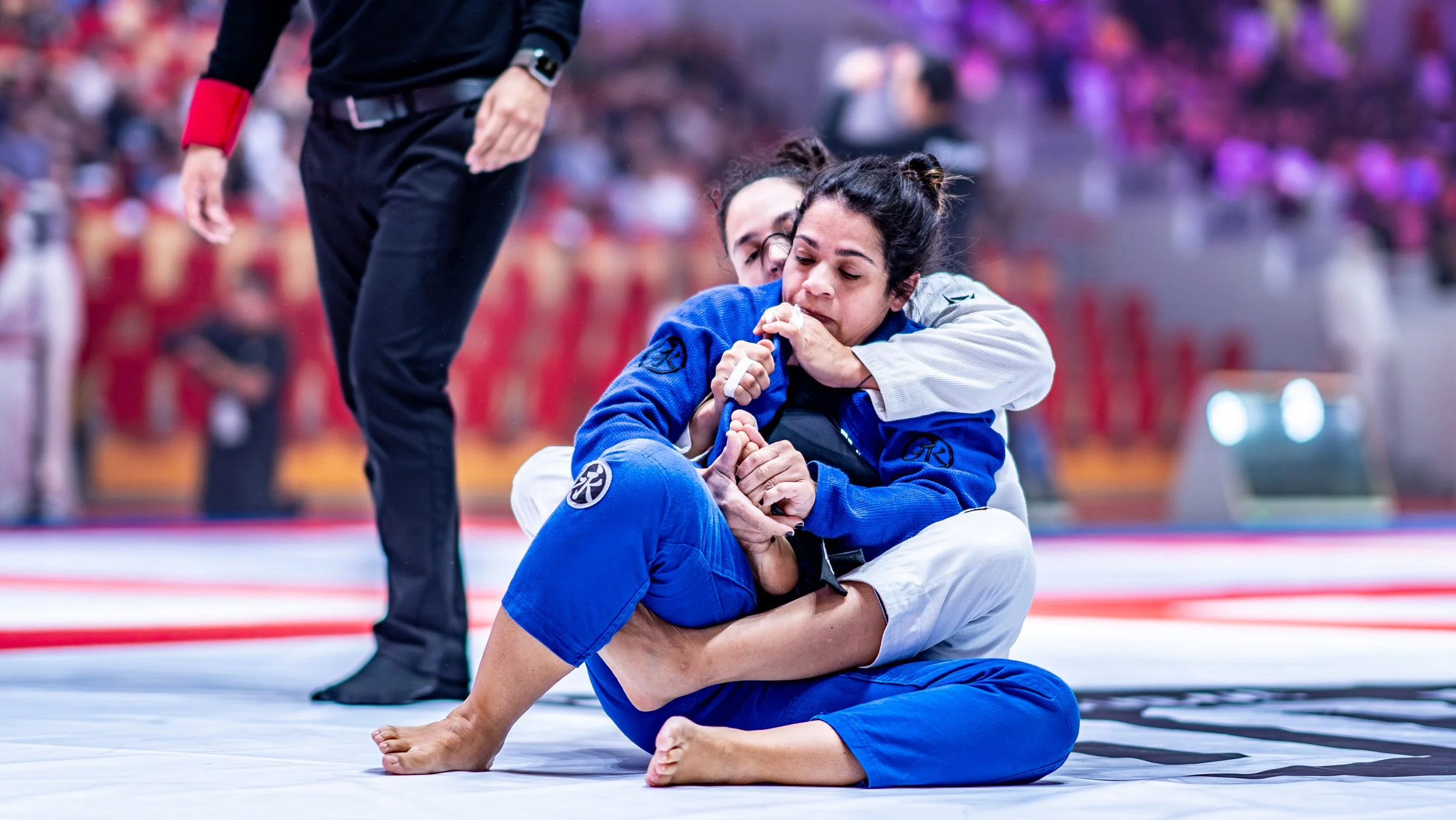 Two women competing in a Brazilian Jiu-Jitsu match, one in a blue gi and the other in a white gi, on the mat, with one woman applying a choke hold while the other defends. A referee stands nearby.