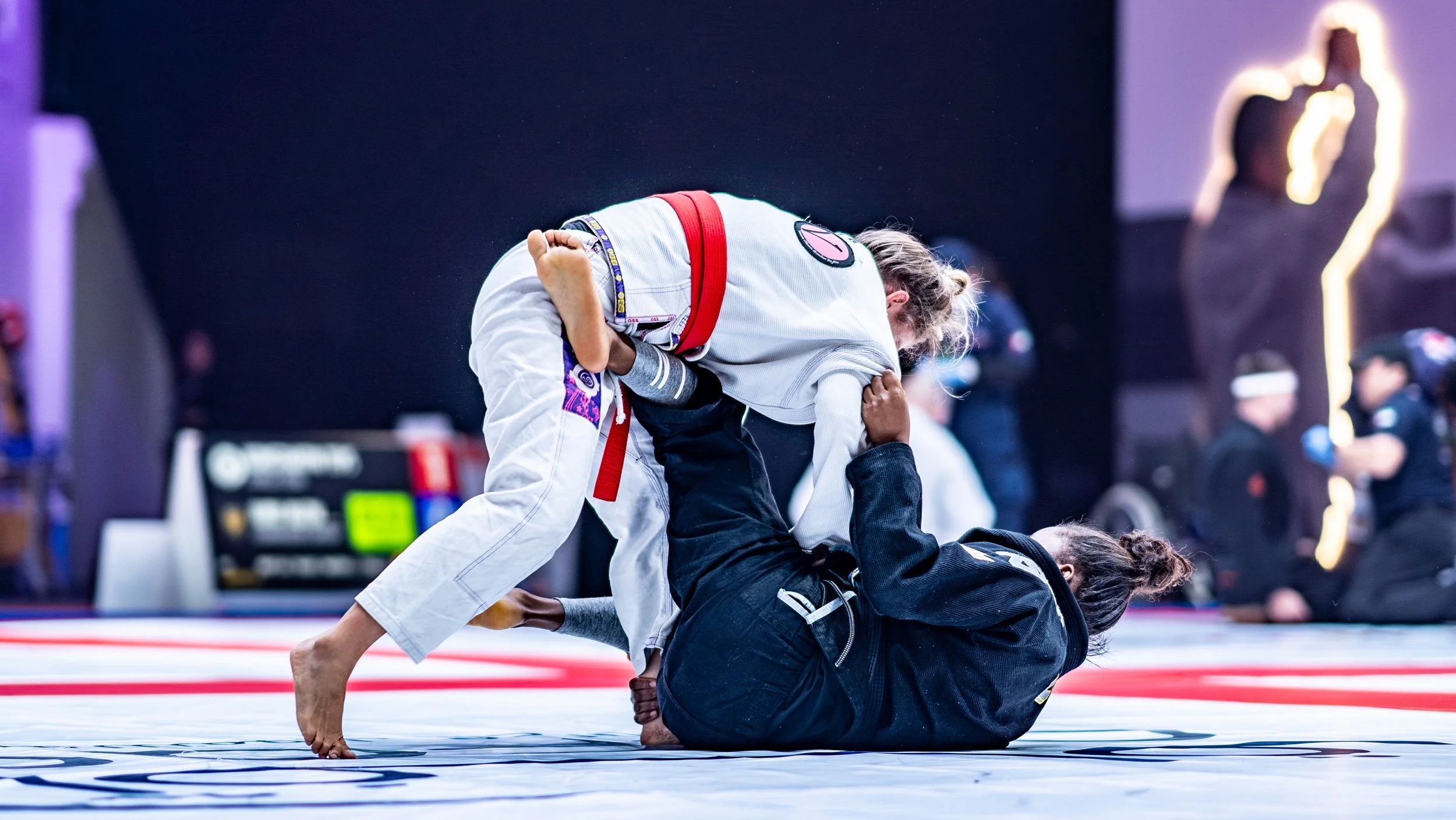 Two female Brazilian Jiu-Jitsu practitioners competing on a mat, one in a white gi and the other in a black gi, with the woman in the black gi on her back and the woman in the white gi leaning over her in a grappling position.