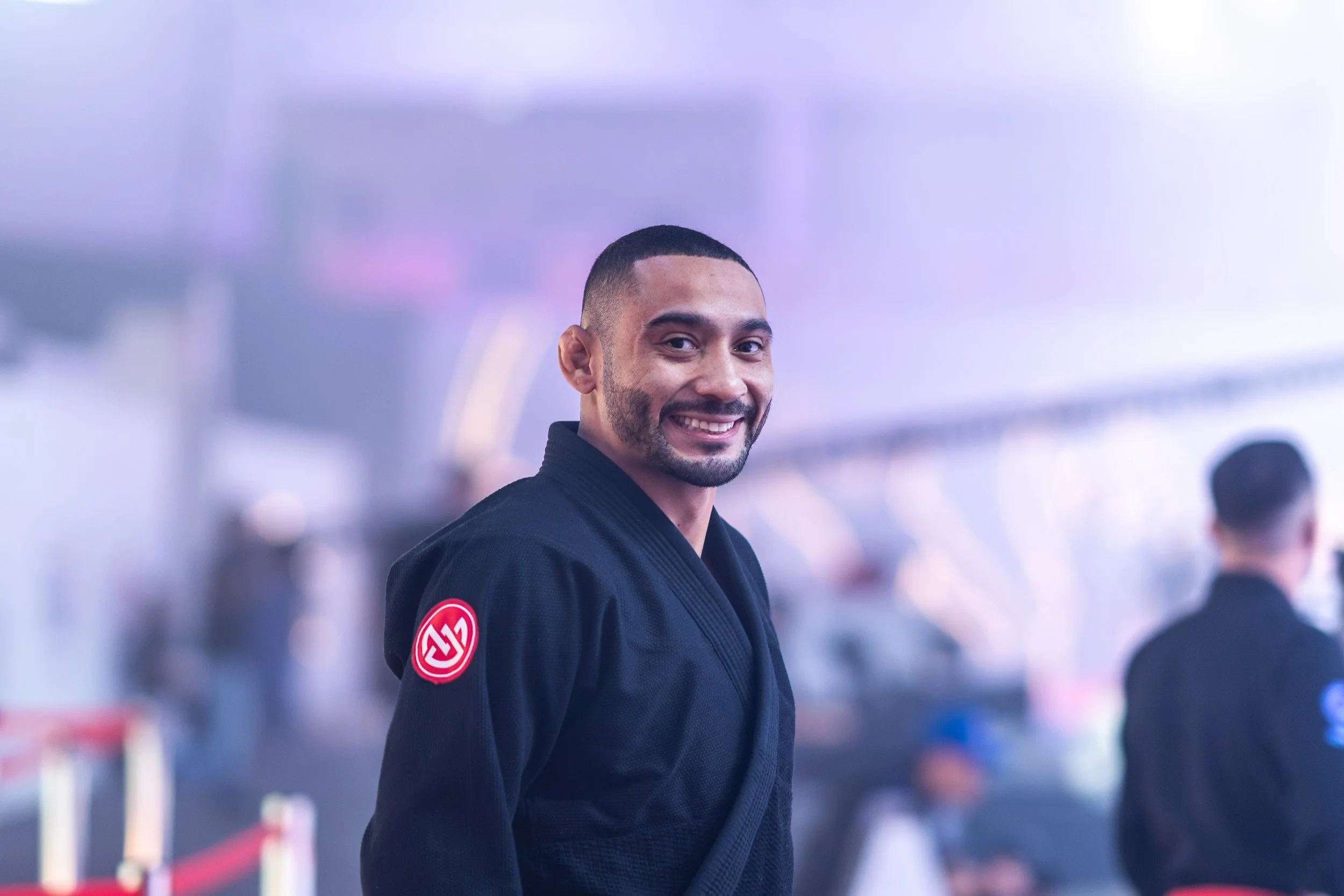 A man smiling in a black martial arts uniform with an AE on the sleeve, standing in a busy indoor setting with other people in the background.