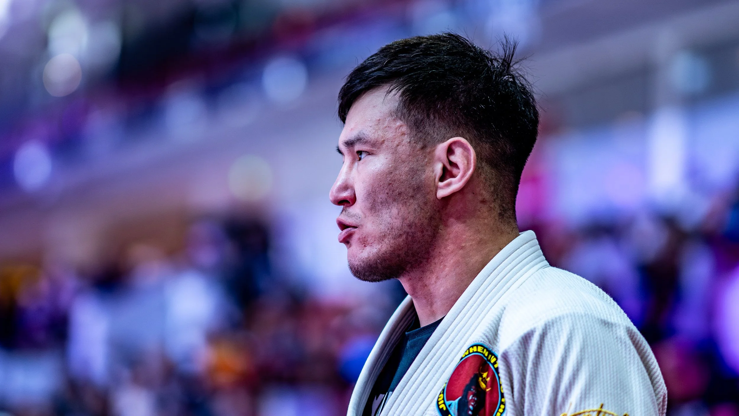 A male Brazilian Jiu-Jitsu athlete in a white gi with a patch on the shoulder, standing in a martial arts arena with a blurred crowd background.