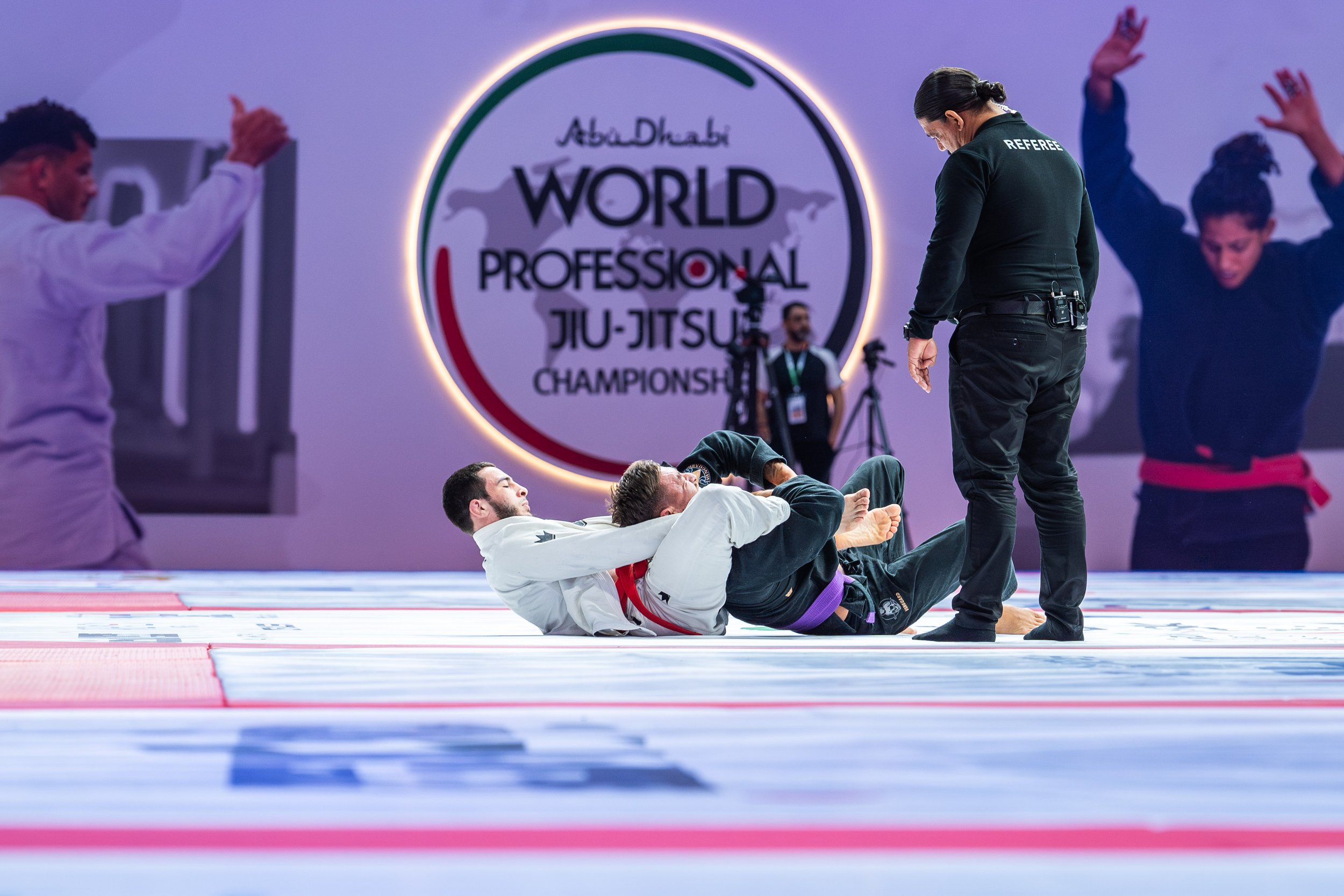 Two male Brazilian Jiu-Jitsu competitors are on the mat during a match at the World Professional Jiu-Jitsu Championship in Abu Dhabi. One competitor in a white gi is on top of a competitor in a black gi, who is lying on his back. A referee, wearing b