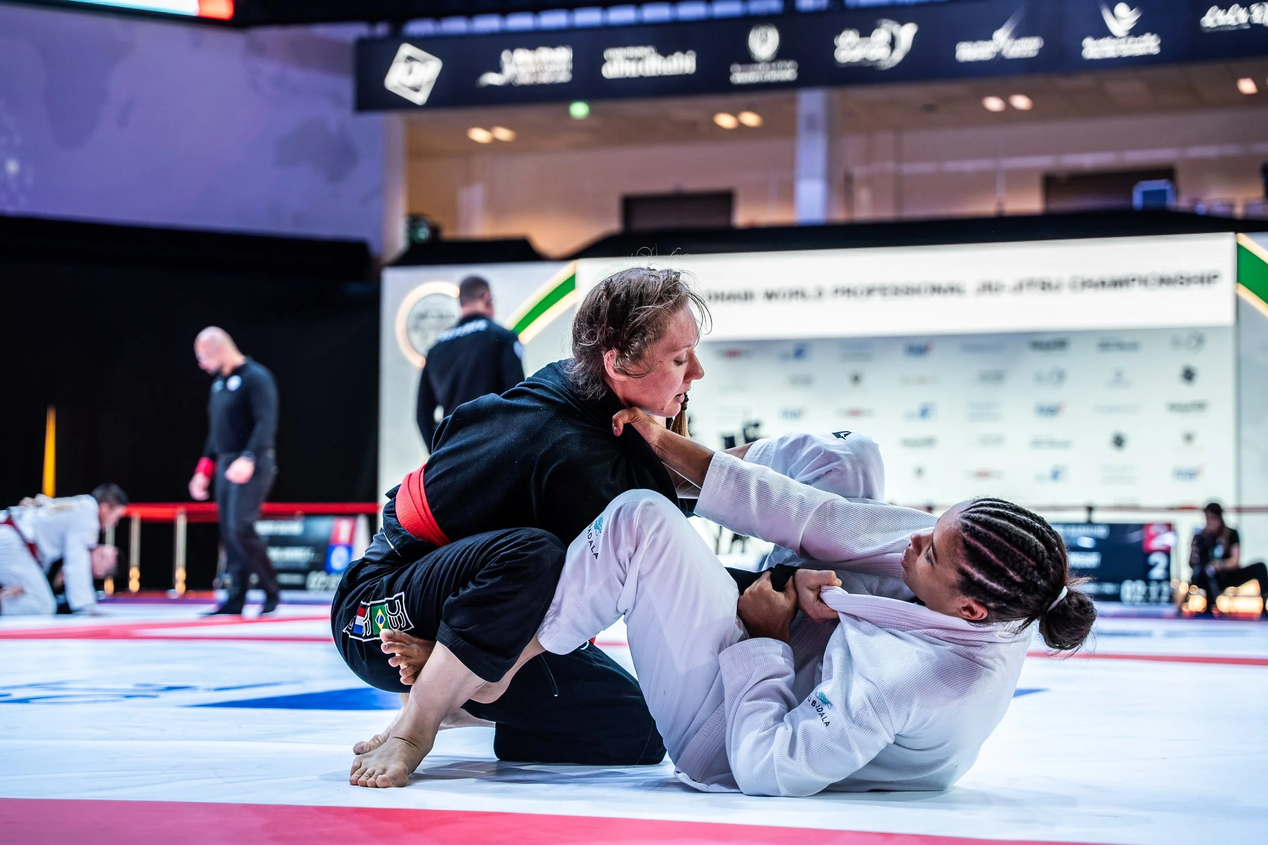 Two women competing in a Brazilian Jiu-Jitsu match on a mat, with one woman in a black gi and the other in a white gi, on a tournament stage with banners and officials in the background.