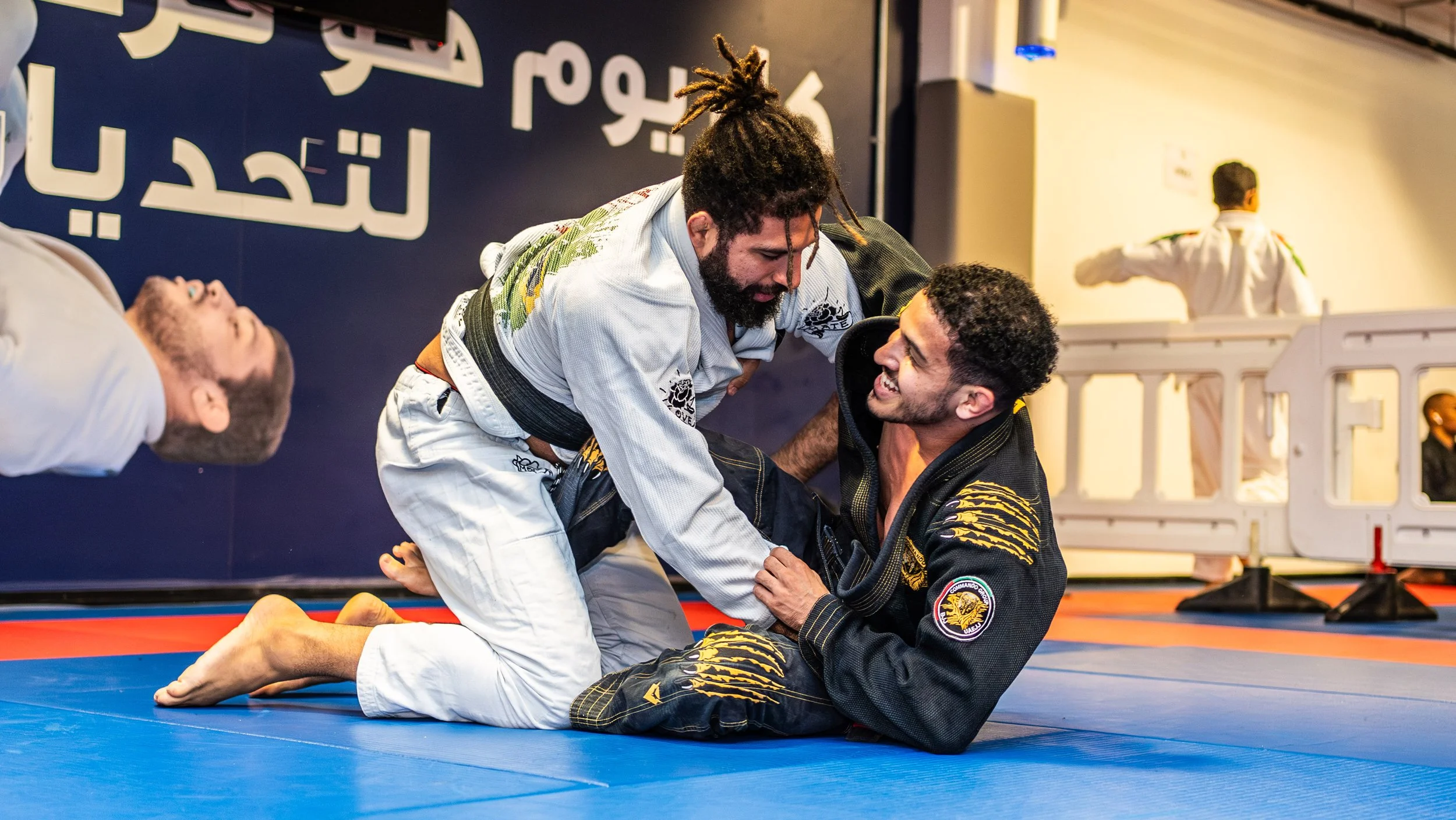 Two men practicing Brazilian Jiu-Jitsu on a blue mat, one in a white gi and the other in a black gi, with a third person lying on the side. They are smiling and engaging in a grappling move, with other practitioners in the background.