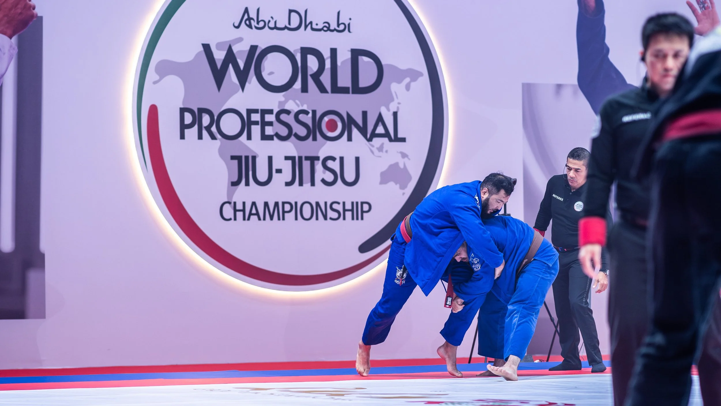 Two competitors in blue gi uniforms practicing jiu-jitsu on the mat, with a referee observing and a large sign in the background reading 'Abu Dhabi World Professional Jiu-Jitsu Championship'.