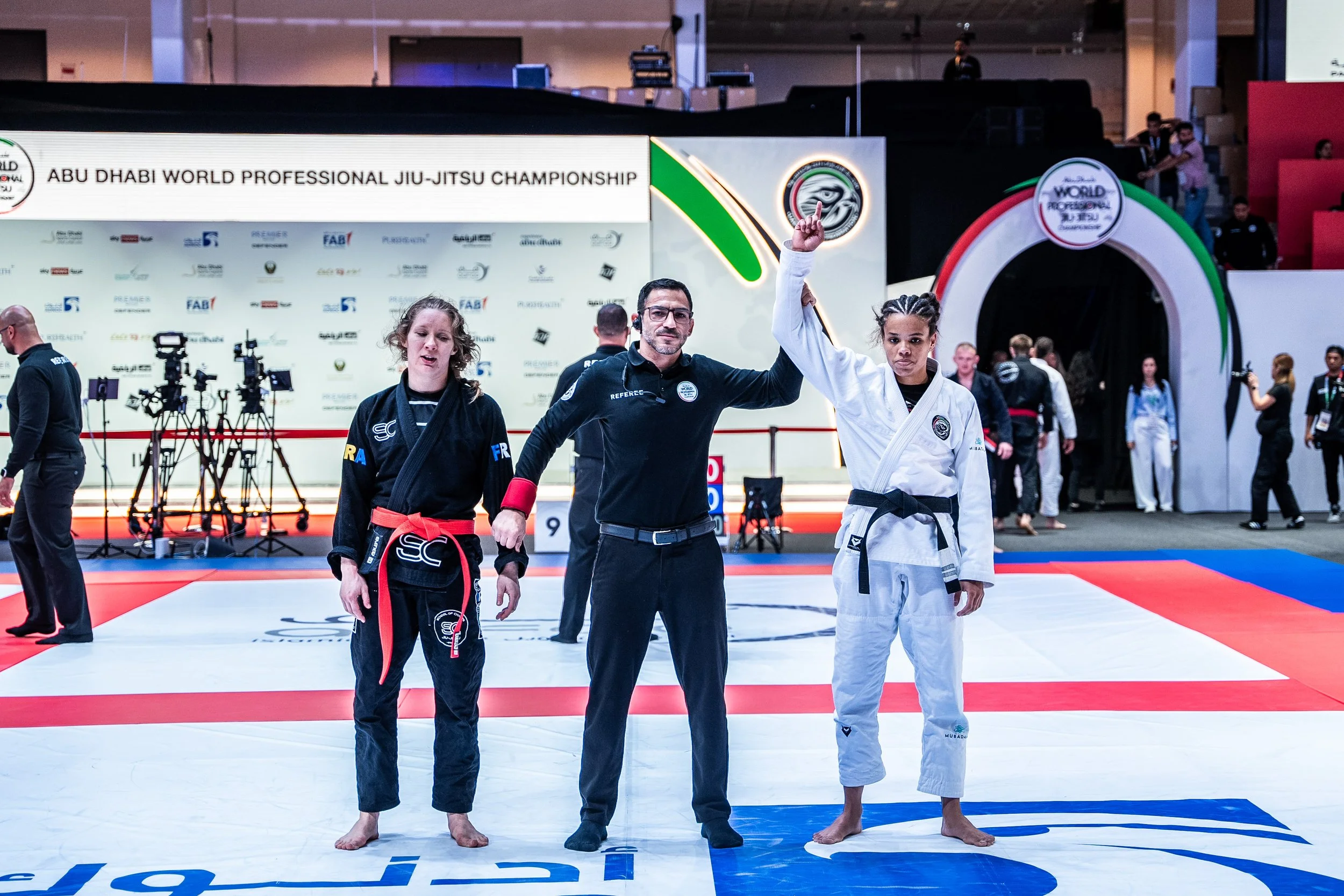 A female judo competitor in a white gi with a black belt standing on a tatami, her arm raised by a referee. Another female competitor in a black gi with a red belt stands beside them. The background shows a banner reading 'Abu Dhabi World Professiona