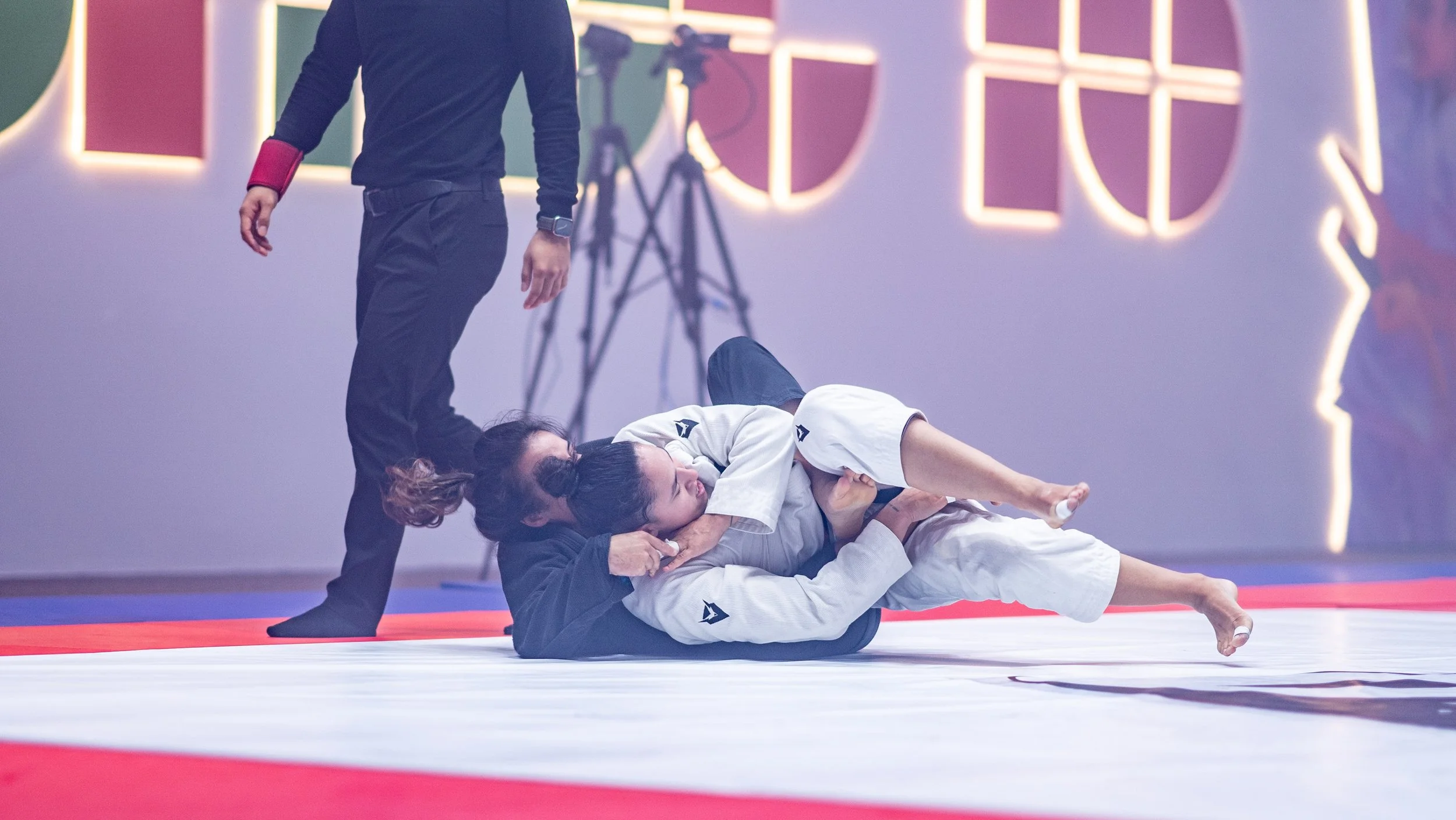 Two women practicing Brazilian Jiu-Jitsu on a mat, one in a white gi on top of the other in a black gi, with a referee standing nearby. The background features illuminated geometric shapes on the wall.