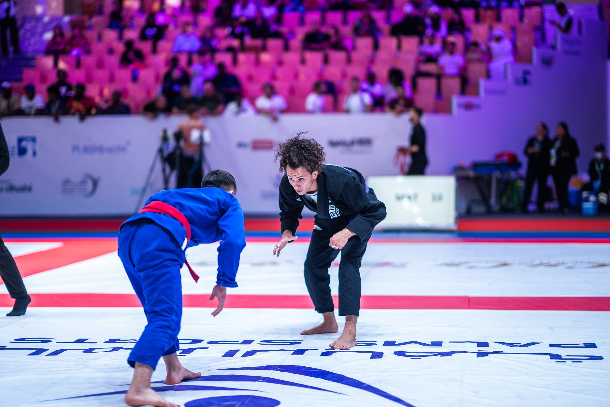Brazilian jiu-jitsu match with two competitors in gi, one in blue and the other in black, facing off on a mat in a large arena, with spectators in the background.