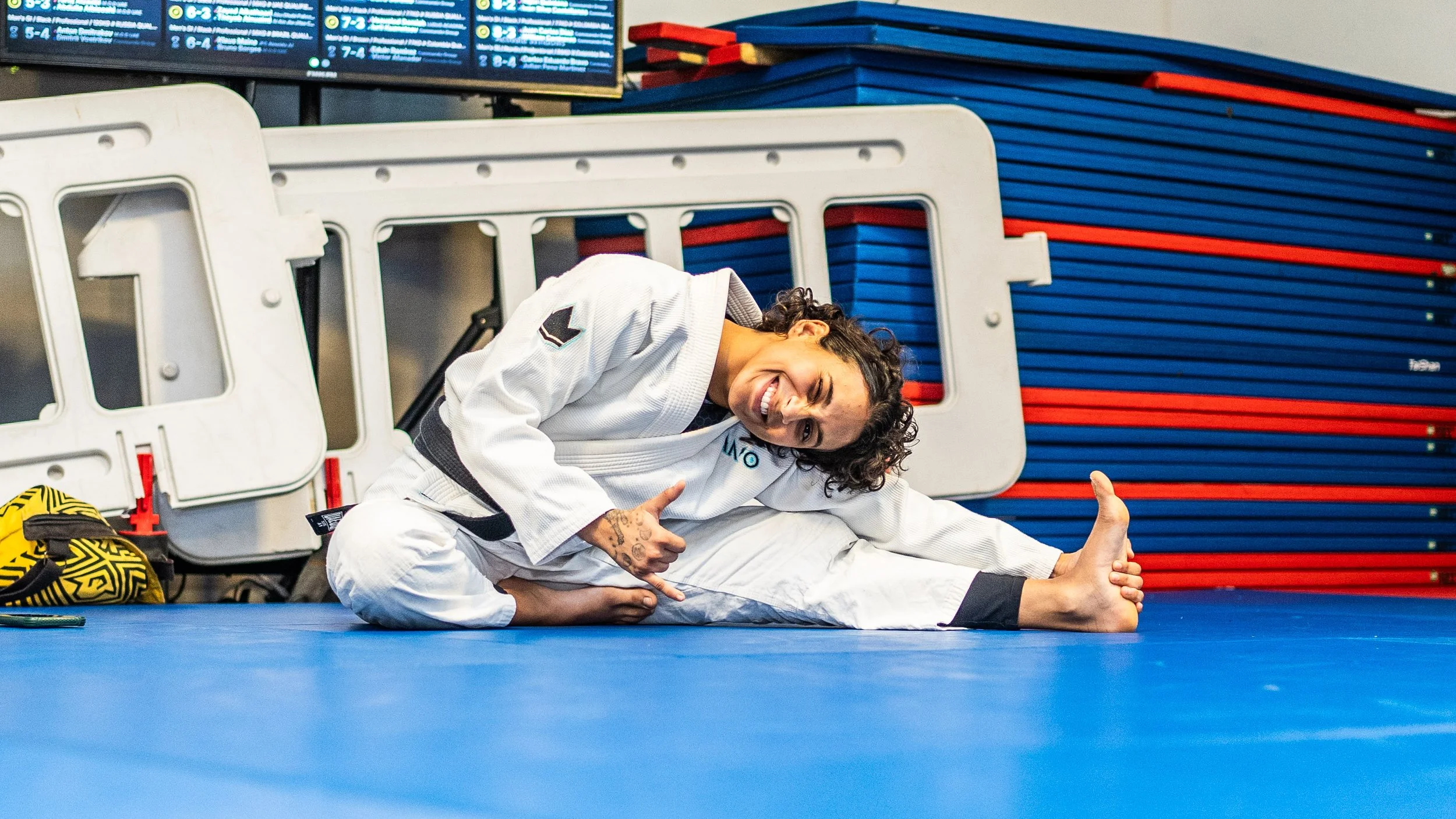 A woman in a white Brazilian Jiu-Jitsu gi smiling and doing a side stretch pose on a blue mat in a martial arts gym, with stacking blue and red mats behind her.