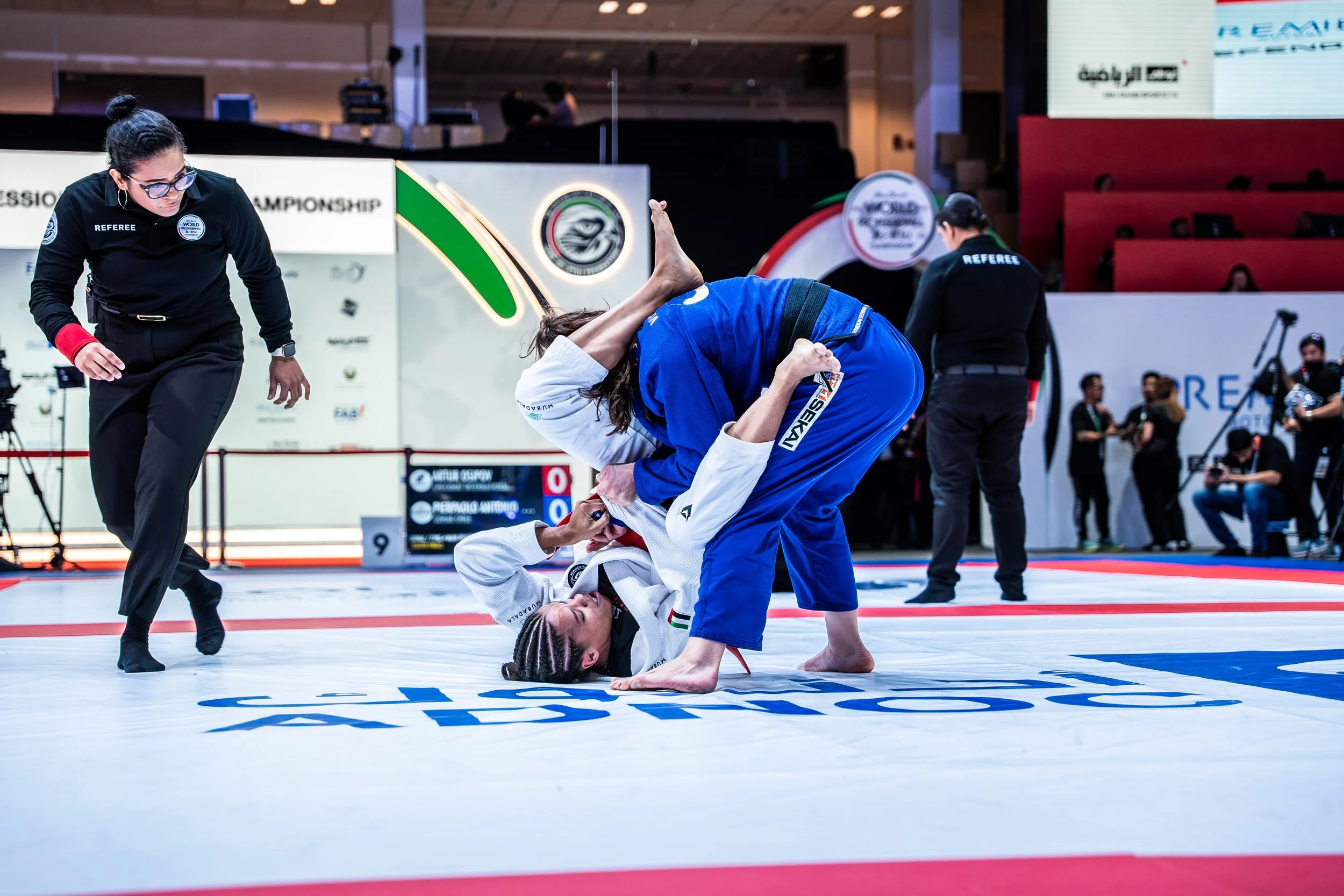 Two female athletes competing in a Brazilian Jiu-Jitsu match, with one in a white gi on her back and the other in a blue gi, pinning her down, under the supervision of a referee, in an indoor sports arena.