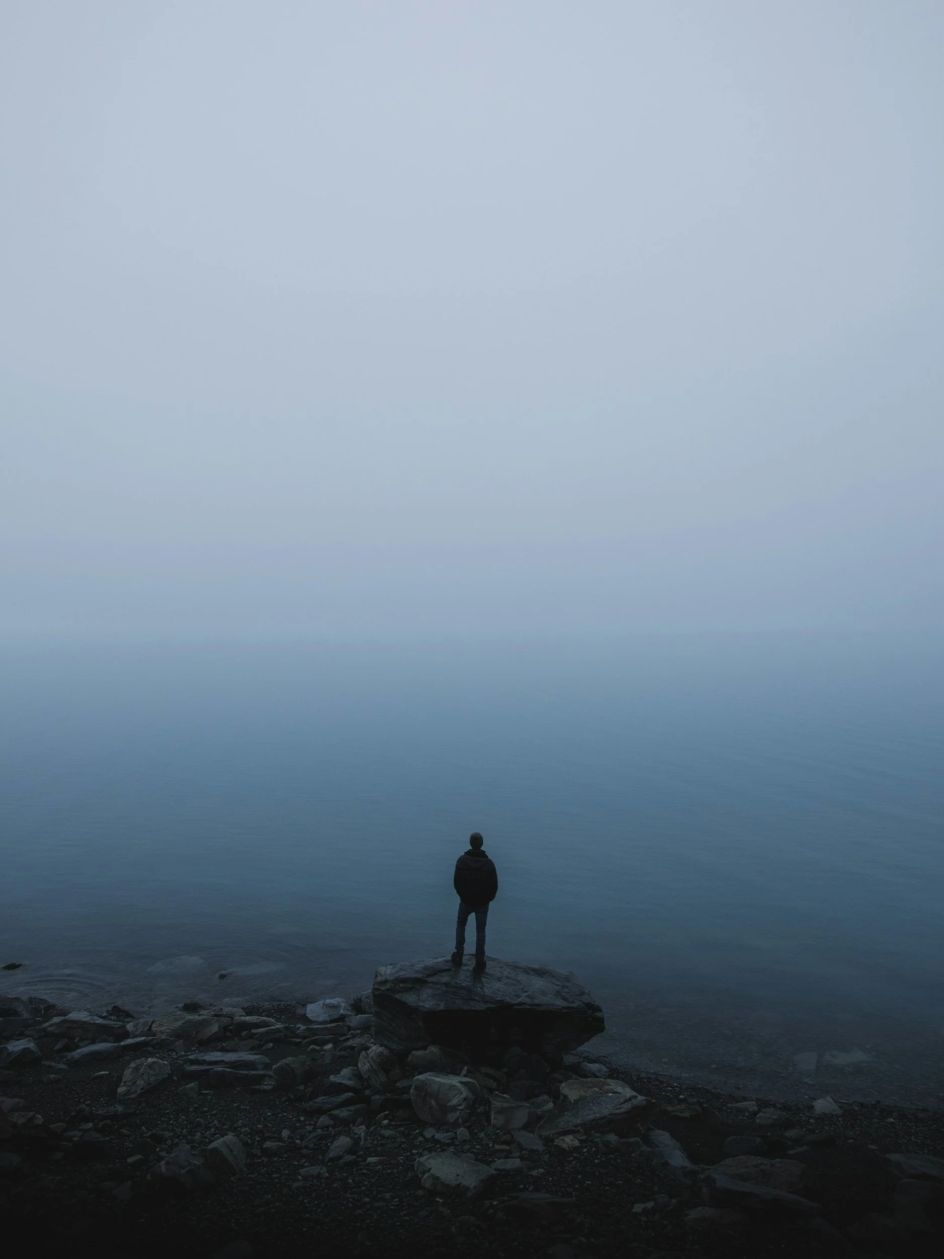 Person standing on rocks at the edge of a calm, expansive body of water, looking out over the horizon under a clear sky.