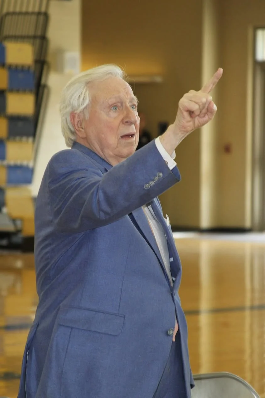 Older man with white hair wearing a blue suit, pointing or gesturing with his right hand in an indoor setting.