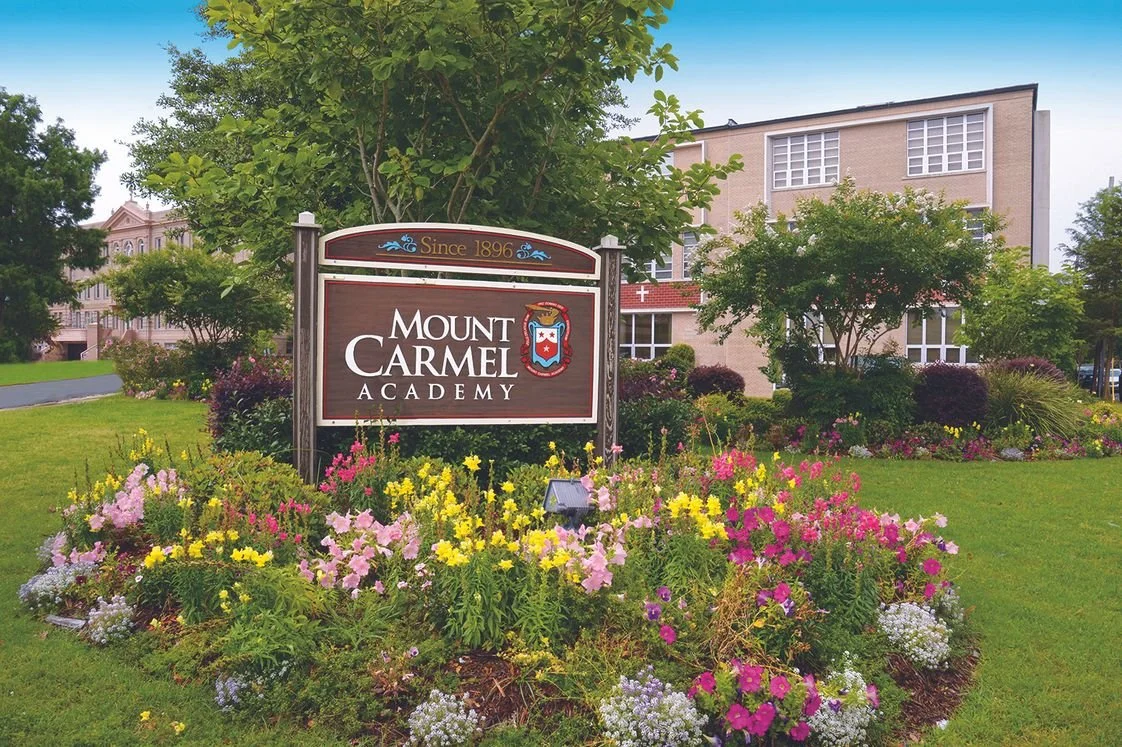 Sign for Mount Carmel Academy surrounded by colorful flowers and greenery, with a school building in the background.