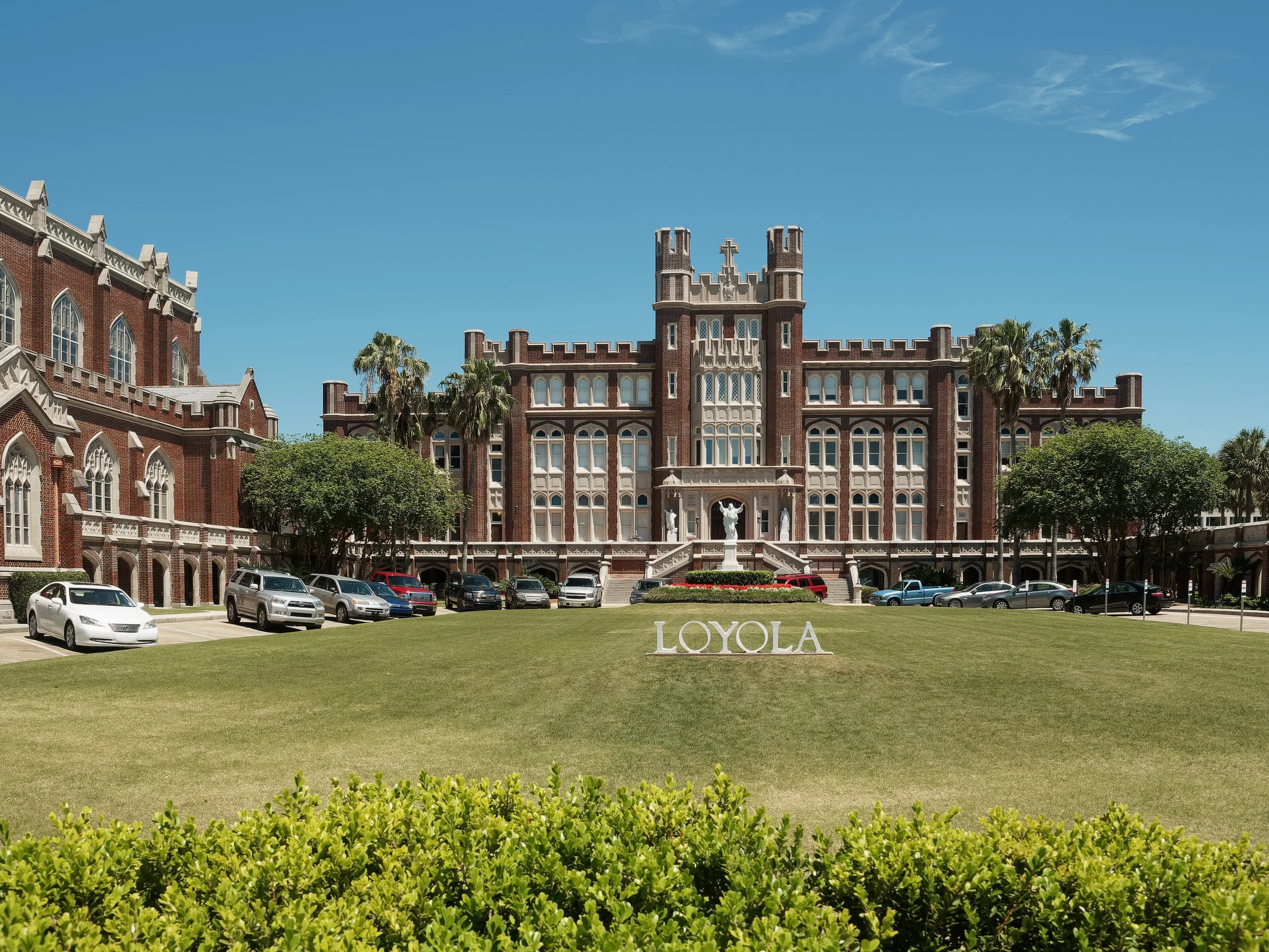 Loyola University campus with historic brick building, green lawn, bushes, parked cars, and a clear blue sky.