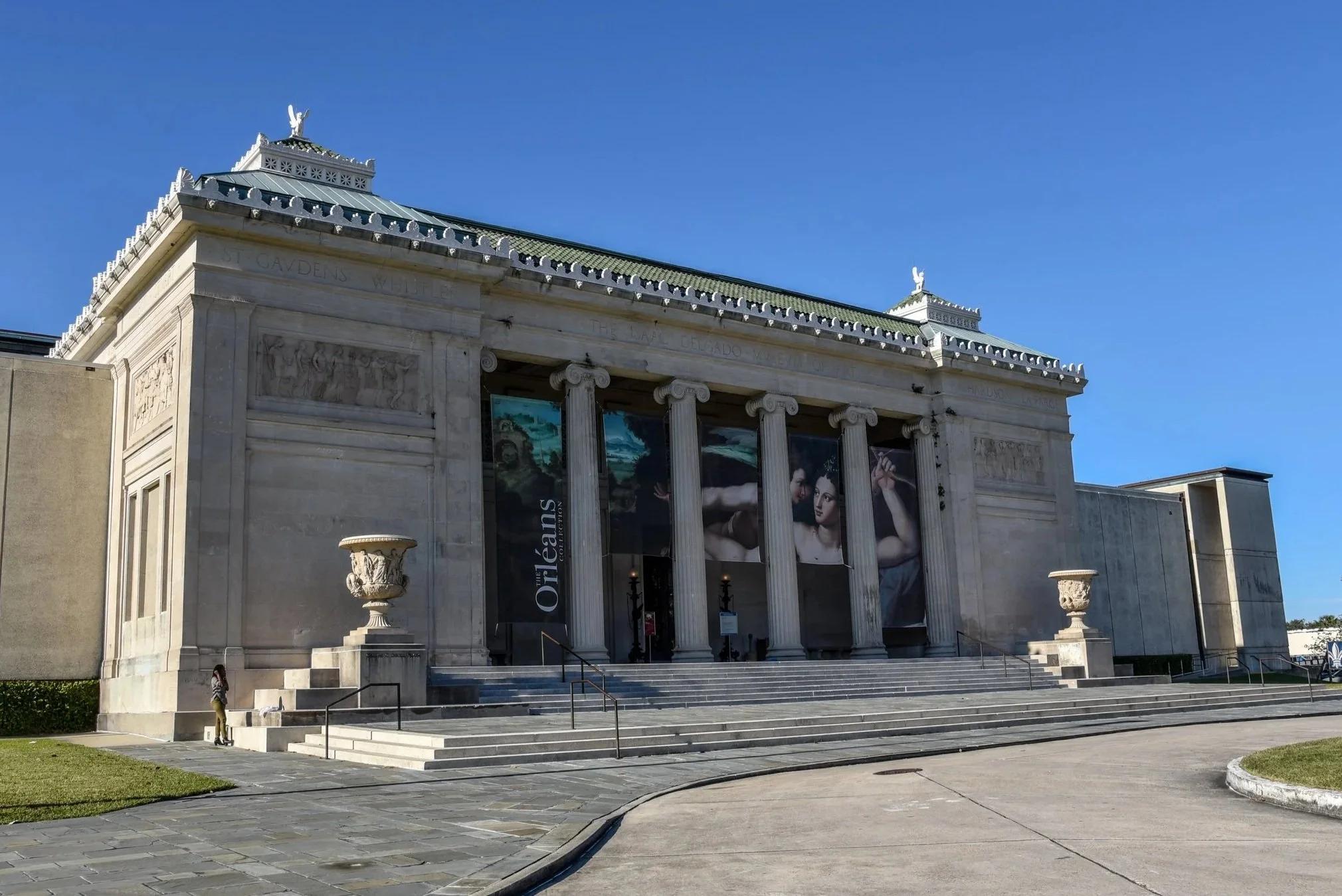 The image shows the front of a classical-style building with columns, steps, and decorative friezes. There is a large poster or banner with a painting of classical figures, and the sky is clear and blue.