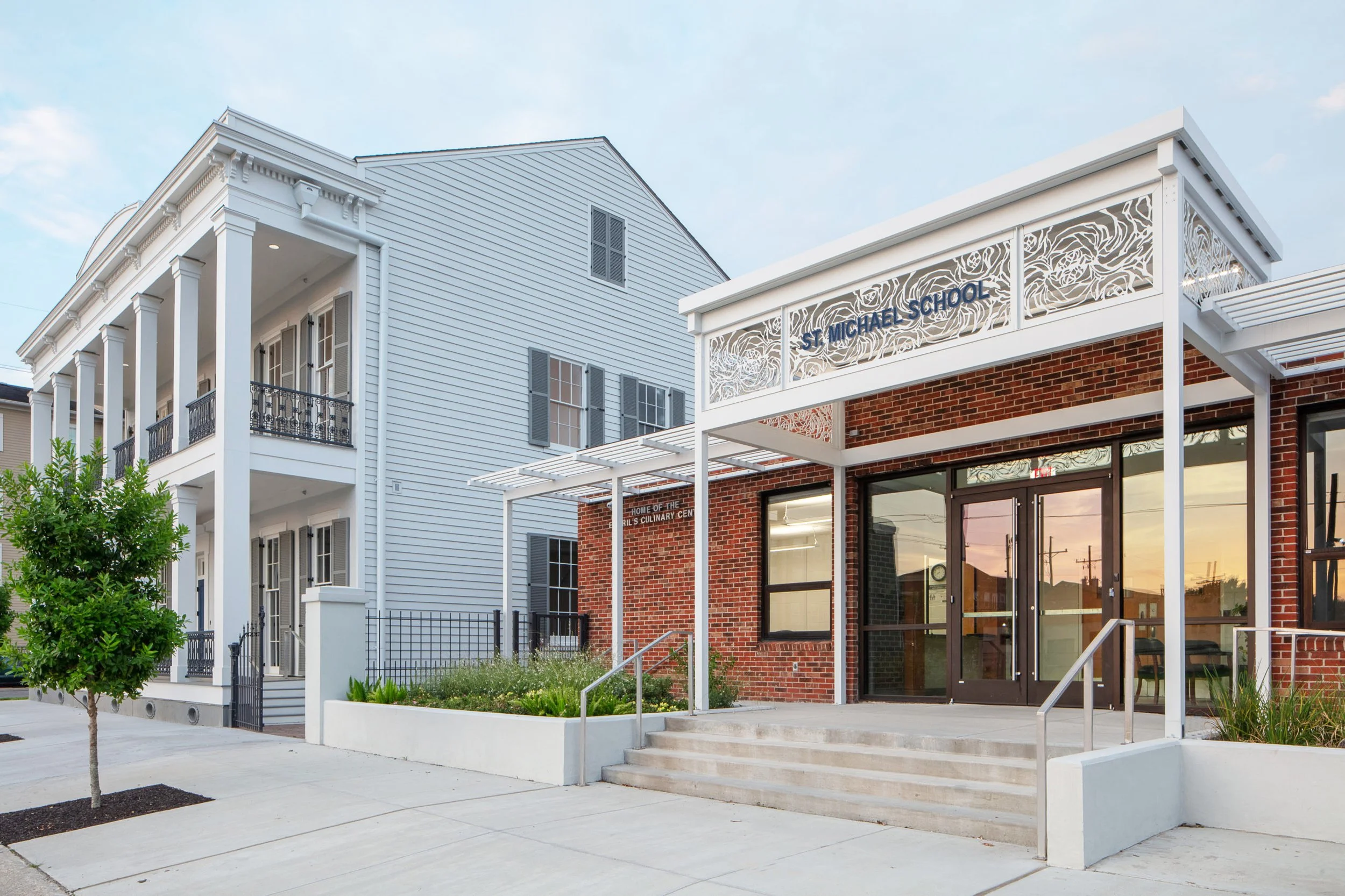 Exterior view of the St. Michael School building, featuring a white wooden structure with columns, black and white railings, a decorative sign with floral patterns, and a red brick entrance, taken during sunset
