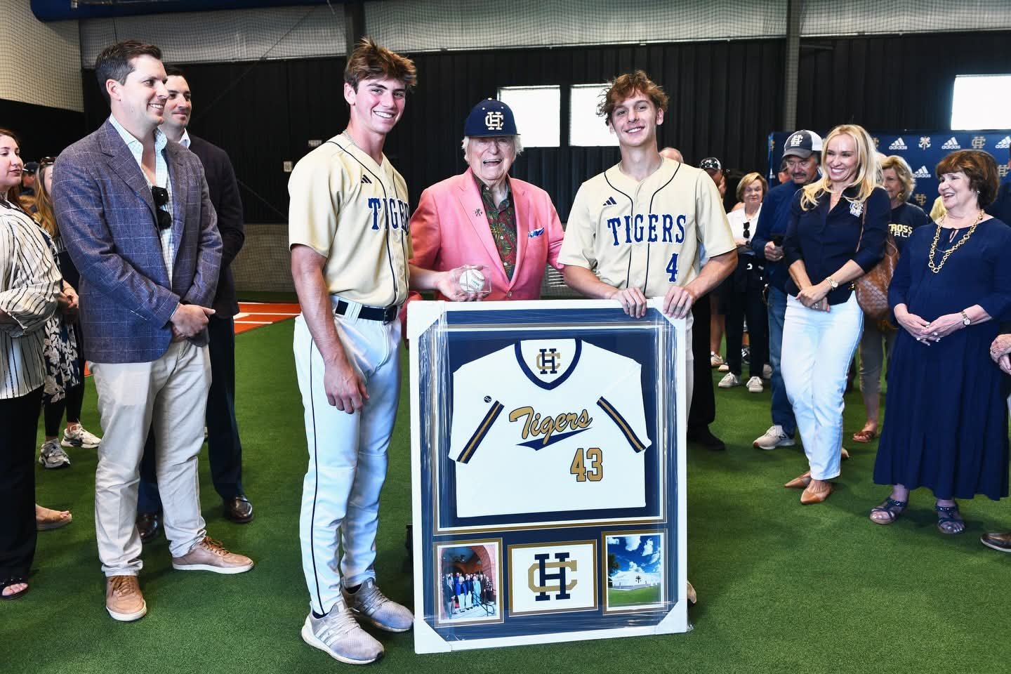 A group of people at a baseball event, including two young baseball players holding a framed jersey with the number 43, an older woman in a pink blazer, and other attendees, some smiling and dressed casually, in an indoor venue with artificial grass.