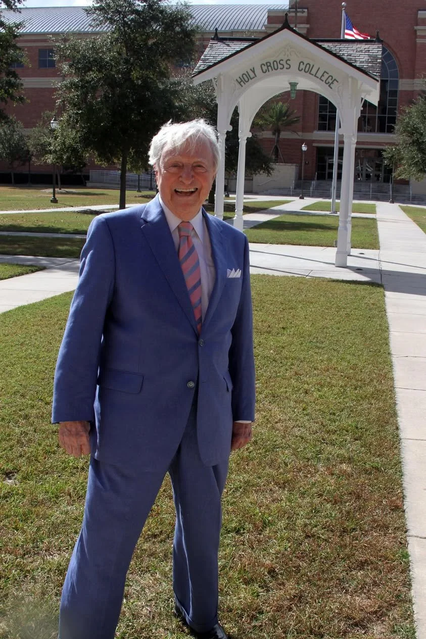An elderly man in a blue suit and pink striped tie standing on a grassy area in front of a white archway that reads 'Holy Cross College,' with buildings and trees in the background.