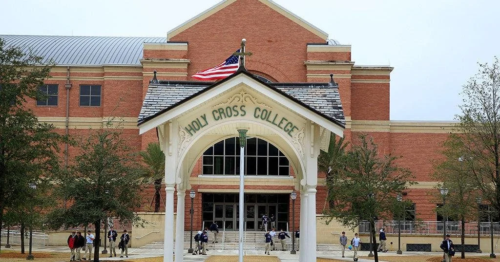 The entrance of Holy Cross College with a white archway, an American flag, and students walking outside.