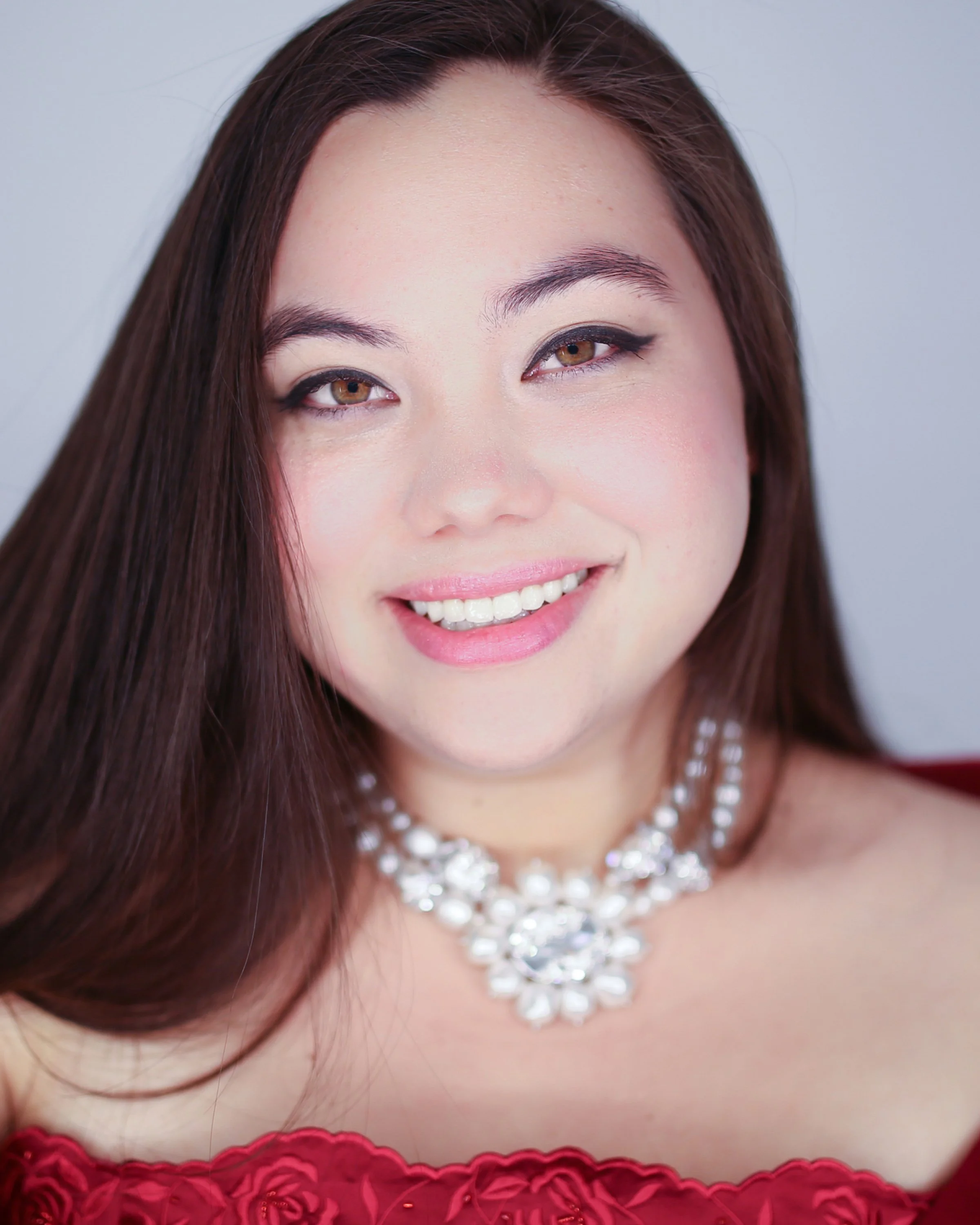 A young woman smiling, wearing a pearl necklace, with long dark hair and a light-colored top.