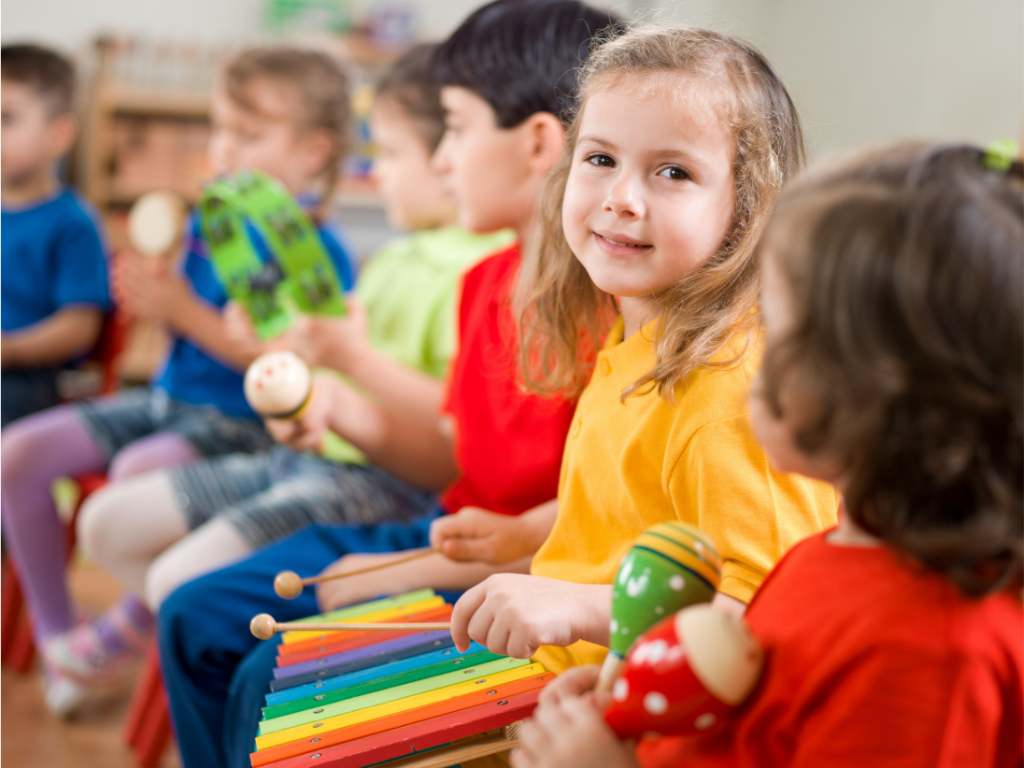 Group of young children sitting in a classroom, holding musical maracas, with a girl in a yellow shirt smiling and looking at the camera, a colorful xylophone in front of her.