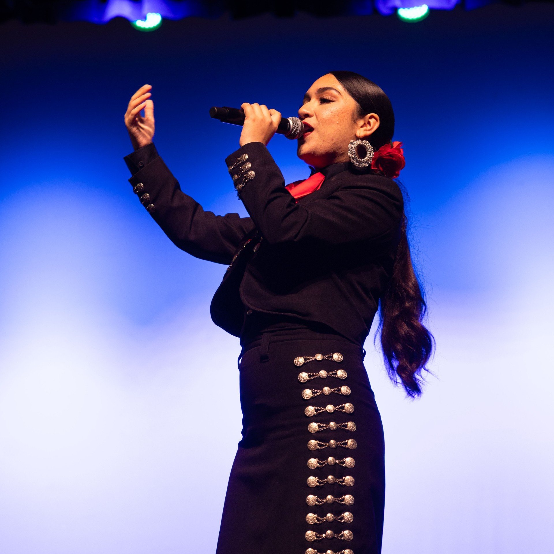 A woman wearing traditional Mexican attire singing into a microphone with a blue background.
