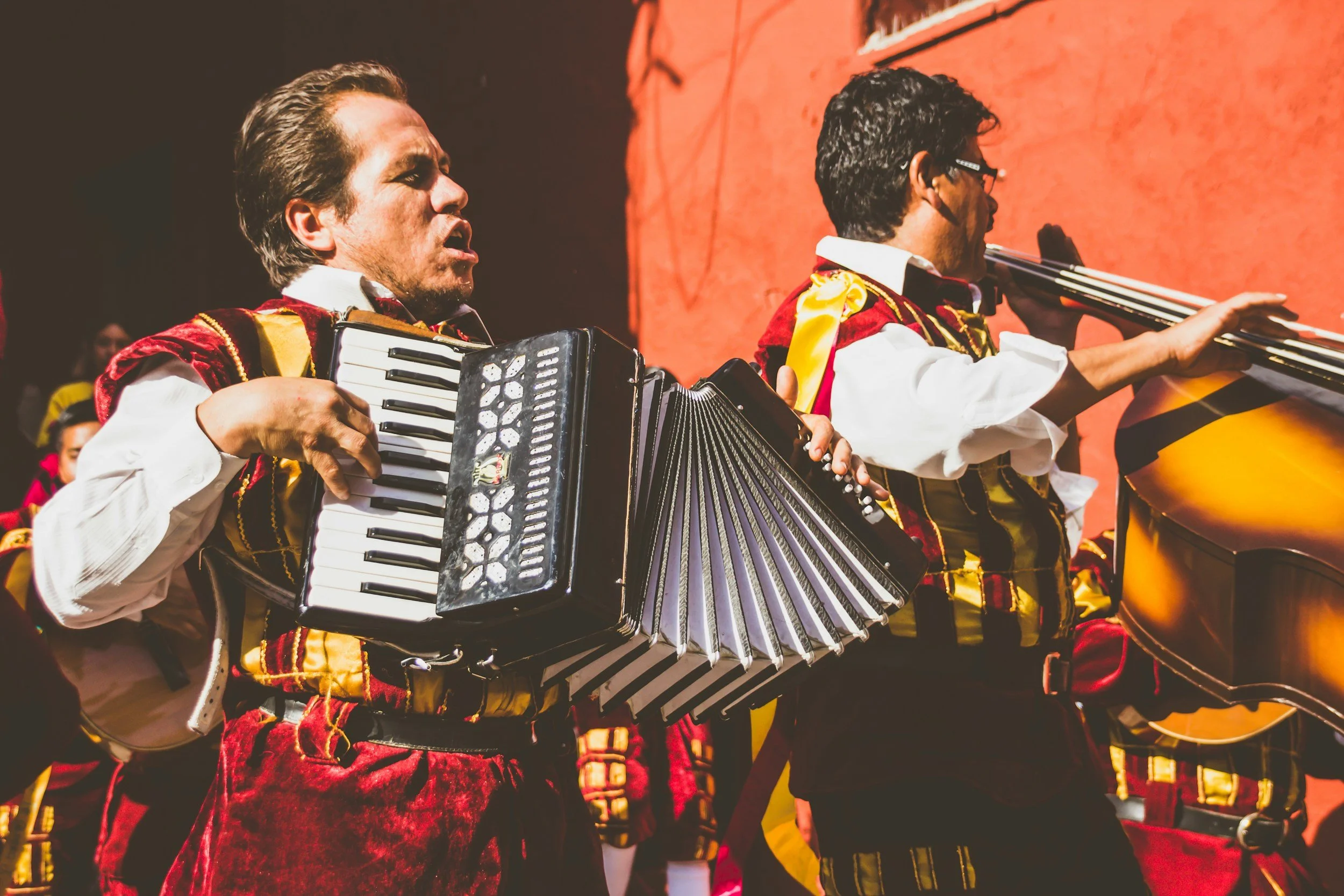 Two men in traditional outfits performing with accordions during a cultural event, with a vivid orange background.