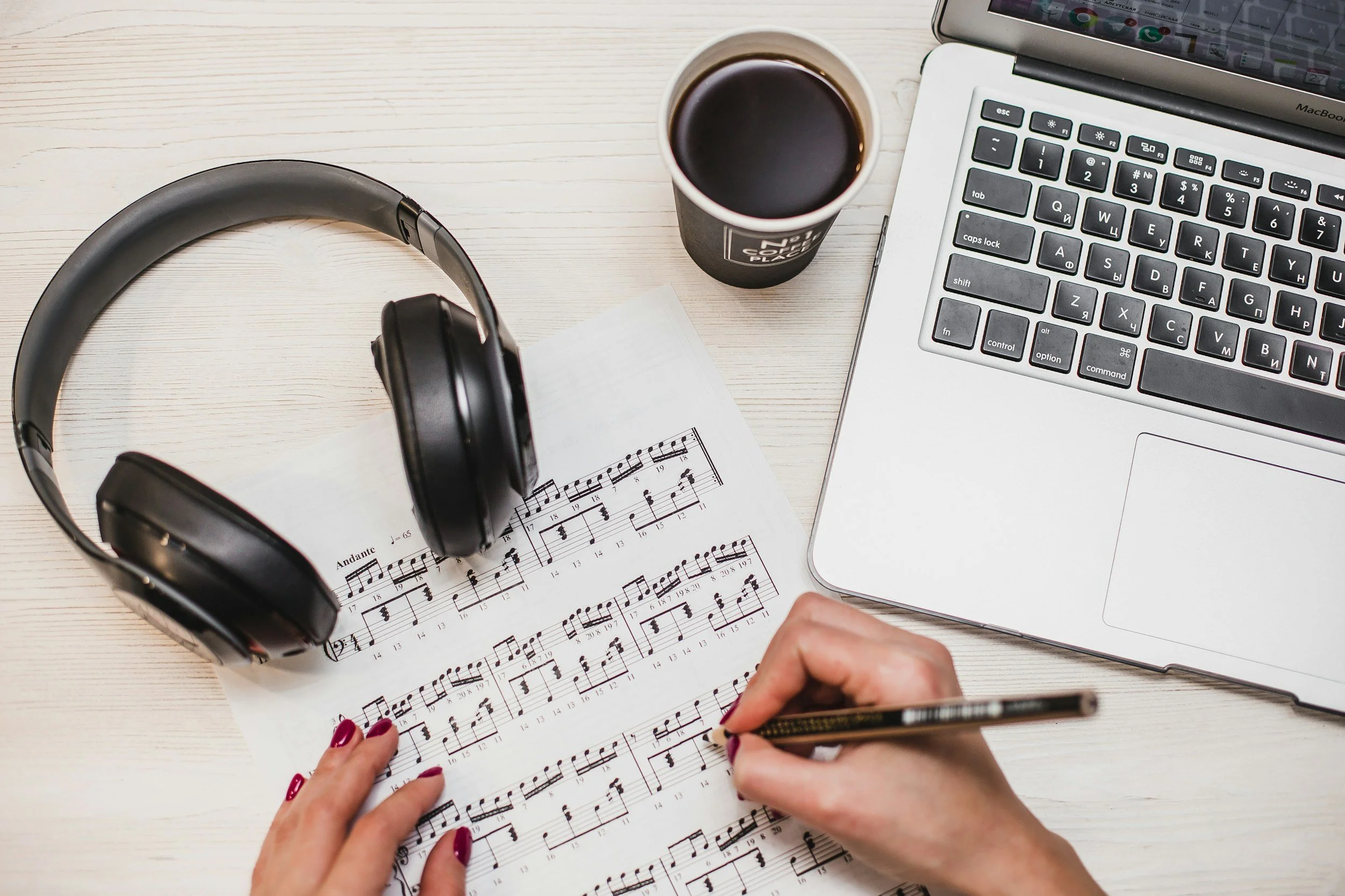 Hands holding a pen and reading sheet music top view, on a desk with a laptop, coffee, headphones, and a smartphone.