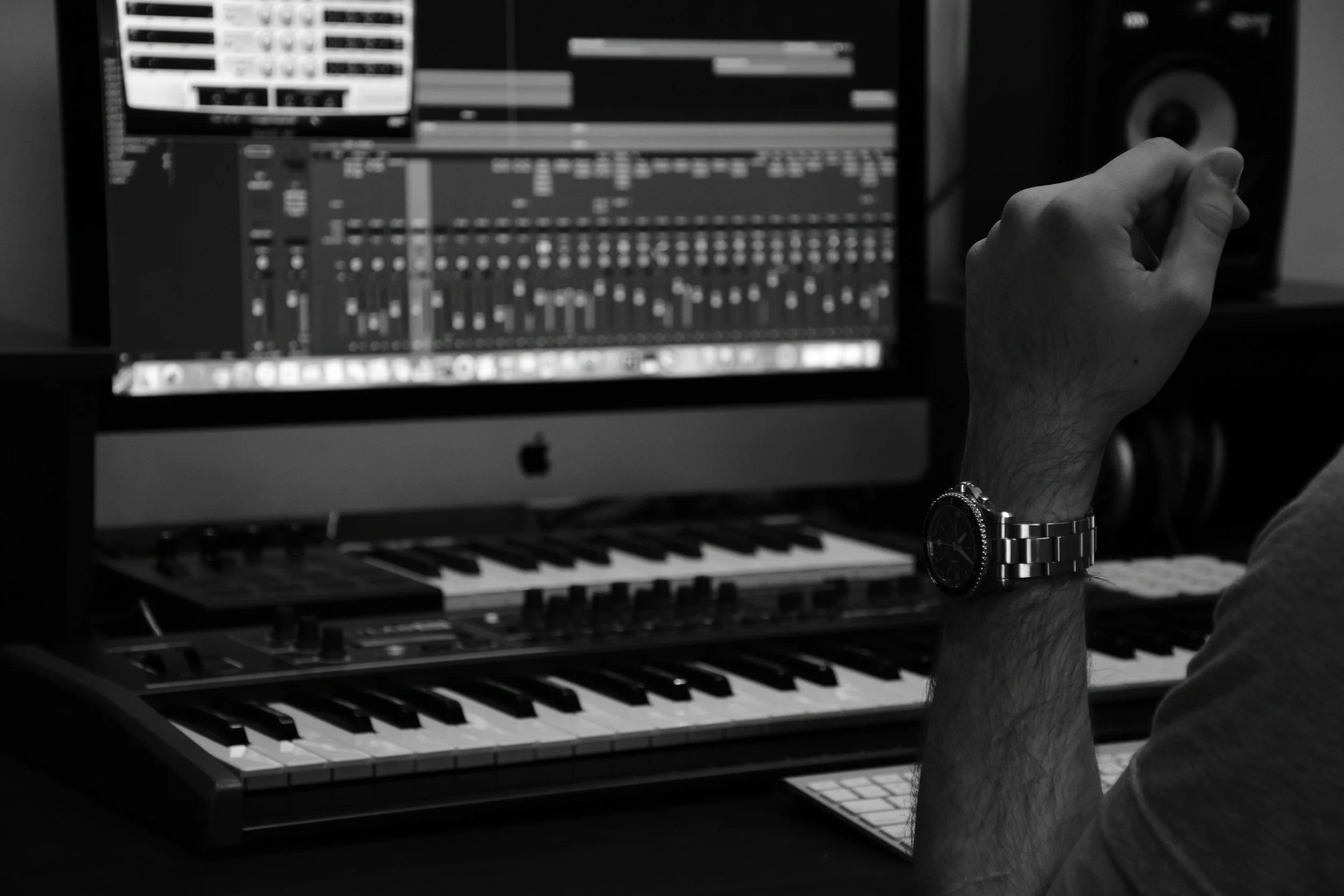 Black and white photo of a person operating music production software on an Apple iMac computer with a digital audio workstation interface displayed. The person's hand is near a speaker, wearing a wristwatch.