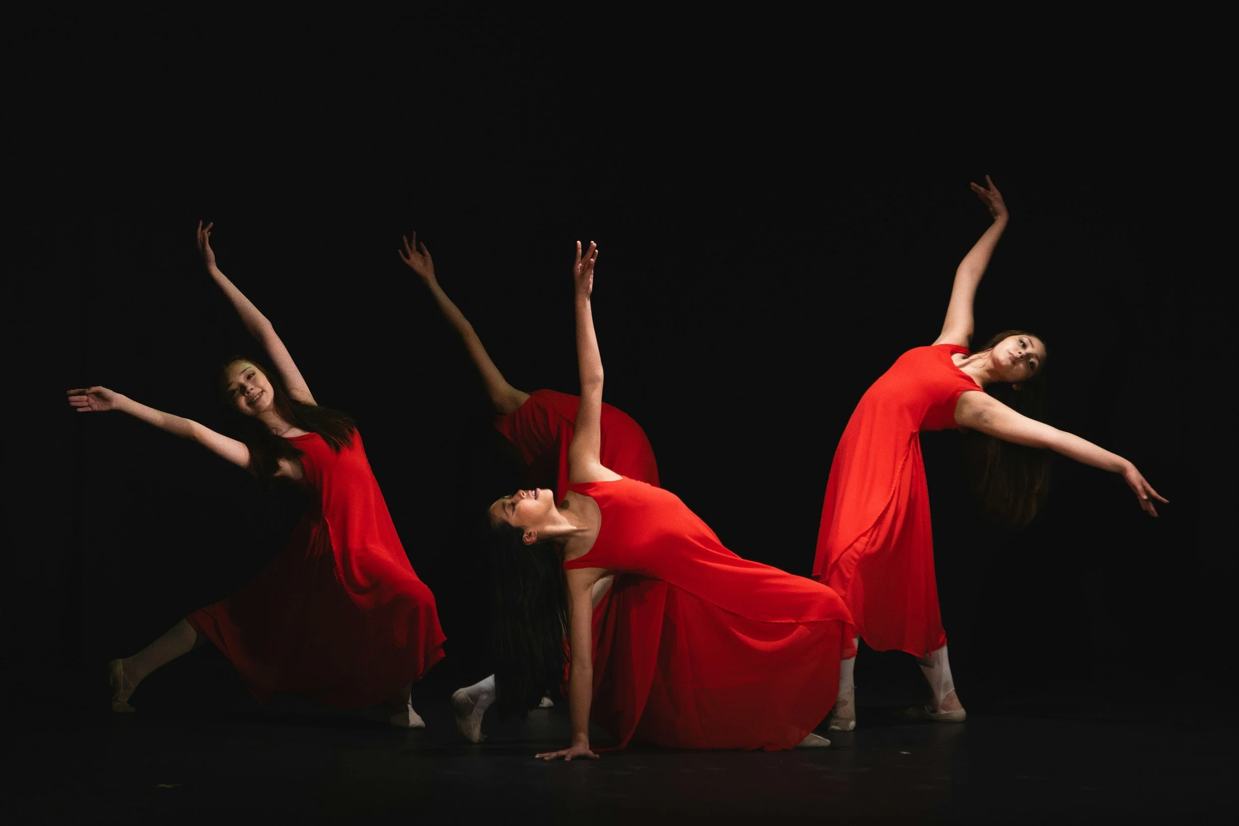 Dance performers in red dresses balancing on one hand with curved bodies and extended arms against a black background.