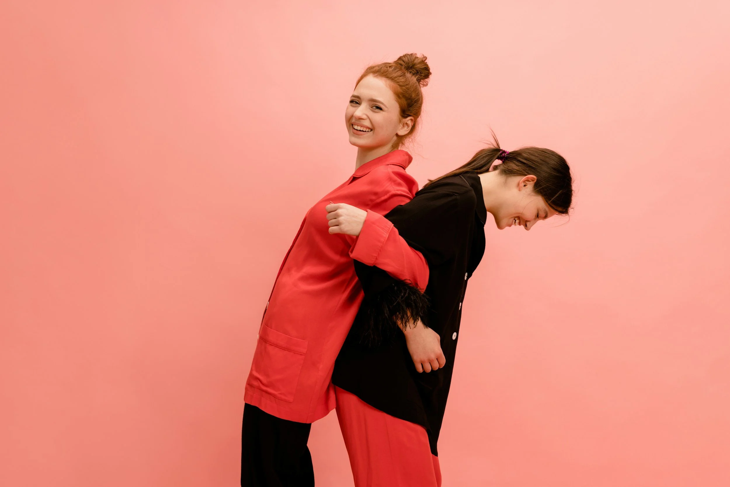 Two women are standing back to back against a pink background. The woman on the left has red hair tied in a bun and is smiling while looking at the camera, wearing a coral-colored shirt. The woman on the right has dark brown hair in a ponytail and is looking down with a smile, wearing a black shirt with rolled-up sleeves.