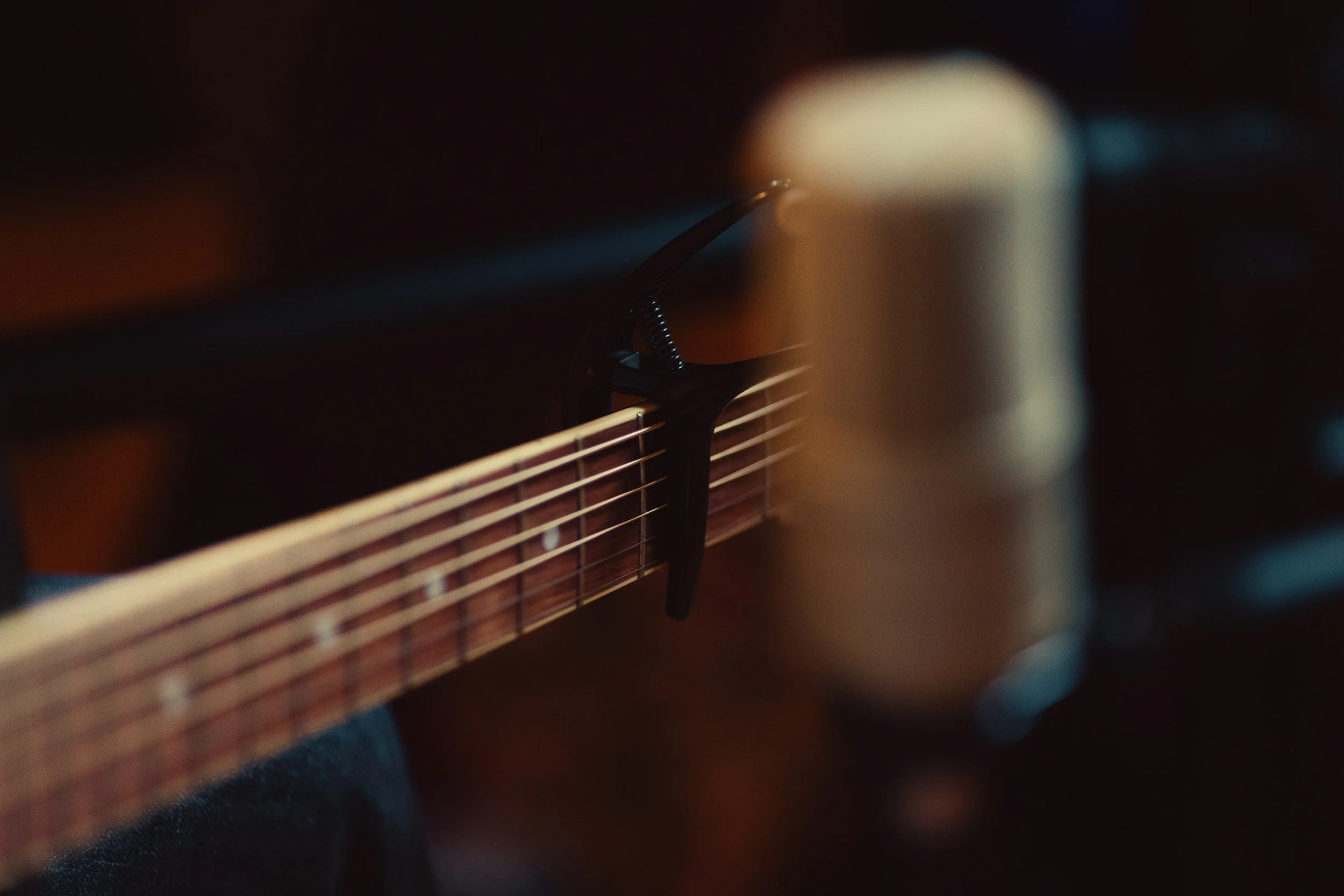 Close-up of the strings and fretboard of an acoustic guitar, with focus on a fret and the side of a finger pressing down on a string, in dim lighting.