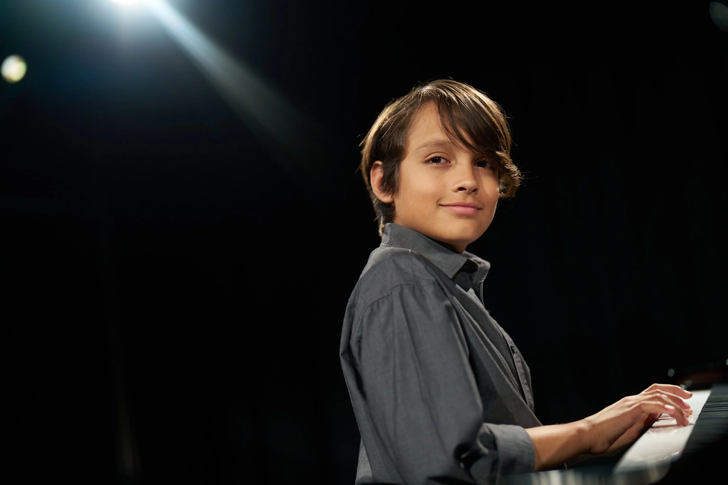 A young boy with brown hair playing the piano on a dark stage, illuminated by overhead stage lights.